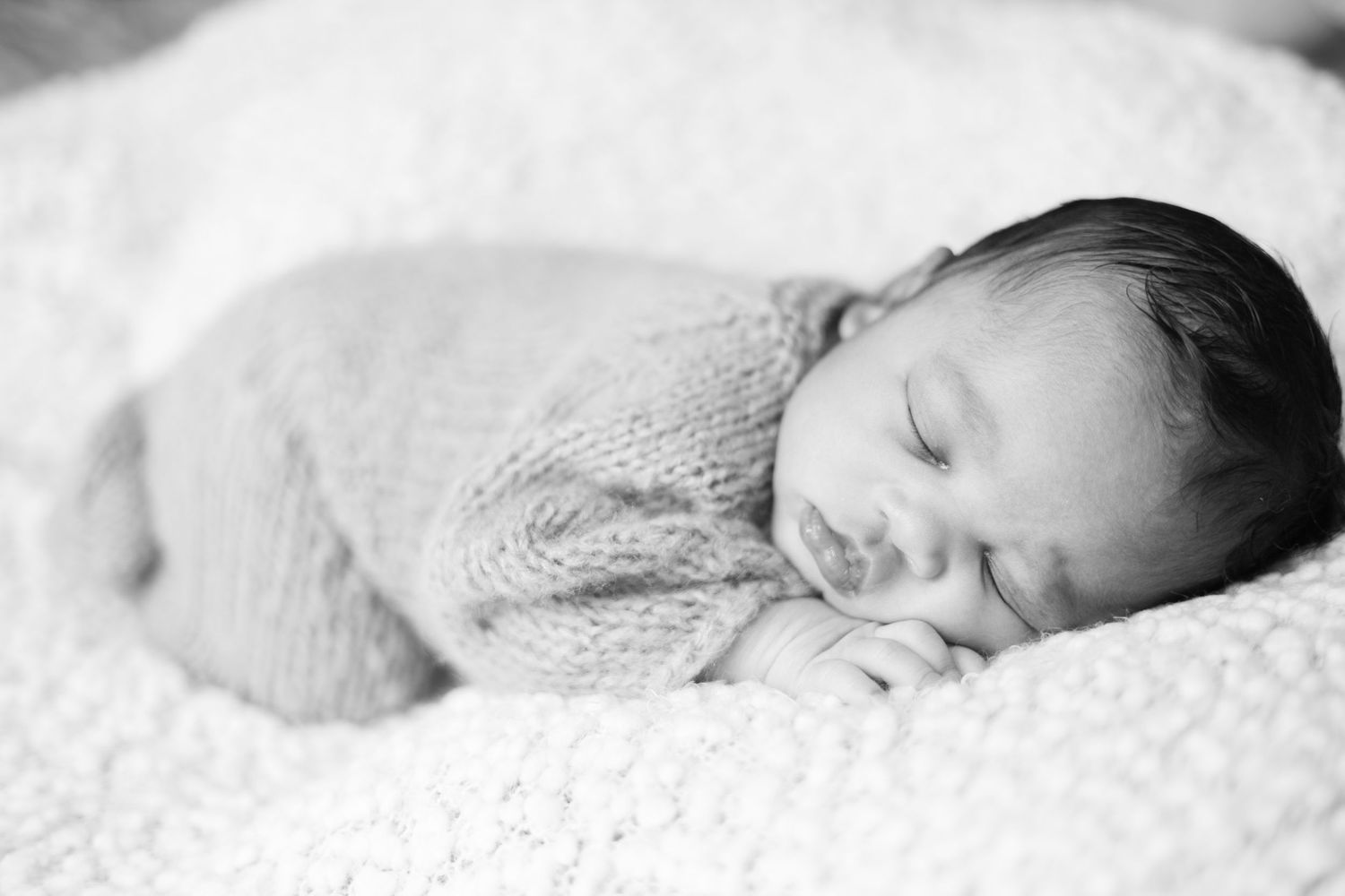 Black and white photo of a sleeping newborn baby wrapped in a soft knit blanket on a fluffy white surface.
