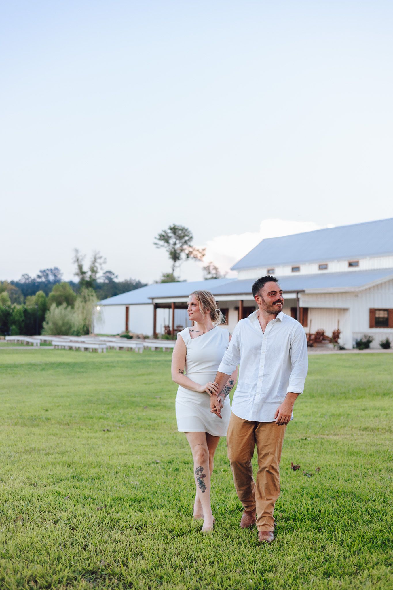 Couple walking hand in hand on green lawn near white barn venue, woman in white dress, man in white shirt and khaki pants.