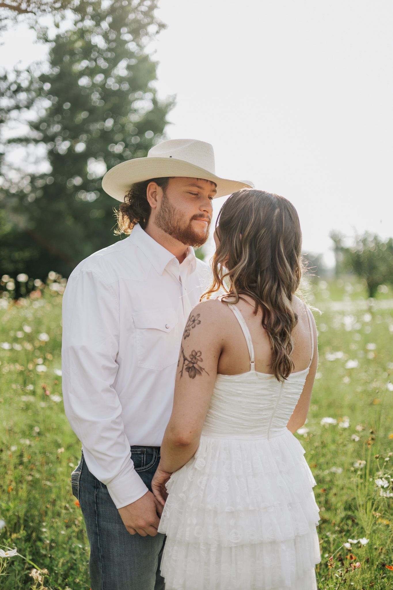 Couple sharing a romantic moment in a wildflower field, man in white shirt and cowboy hat, woman in white dress with floral tattoo.