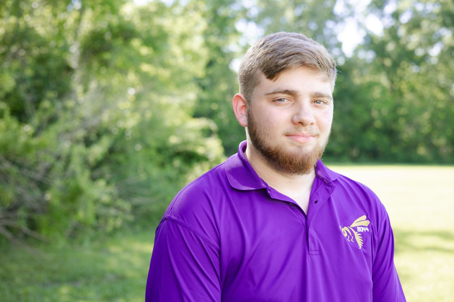 Young man with beard wearing purple polo shirt smiling outdoors with green trees in background.