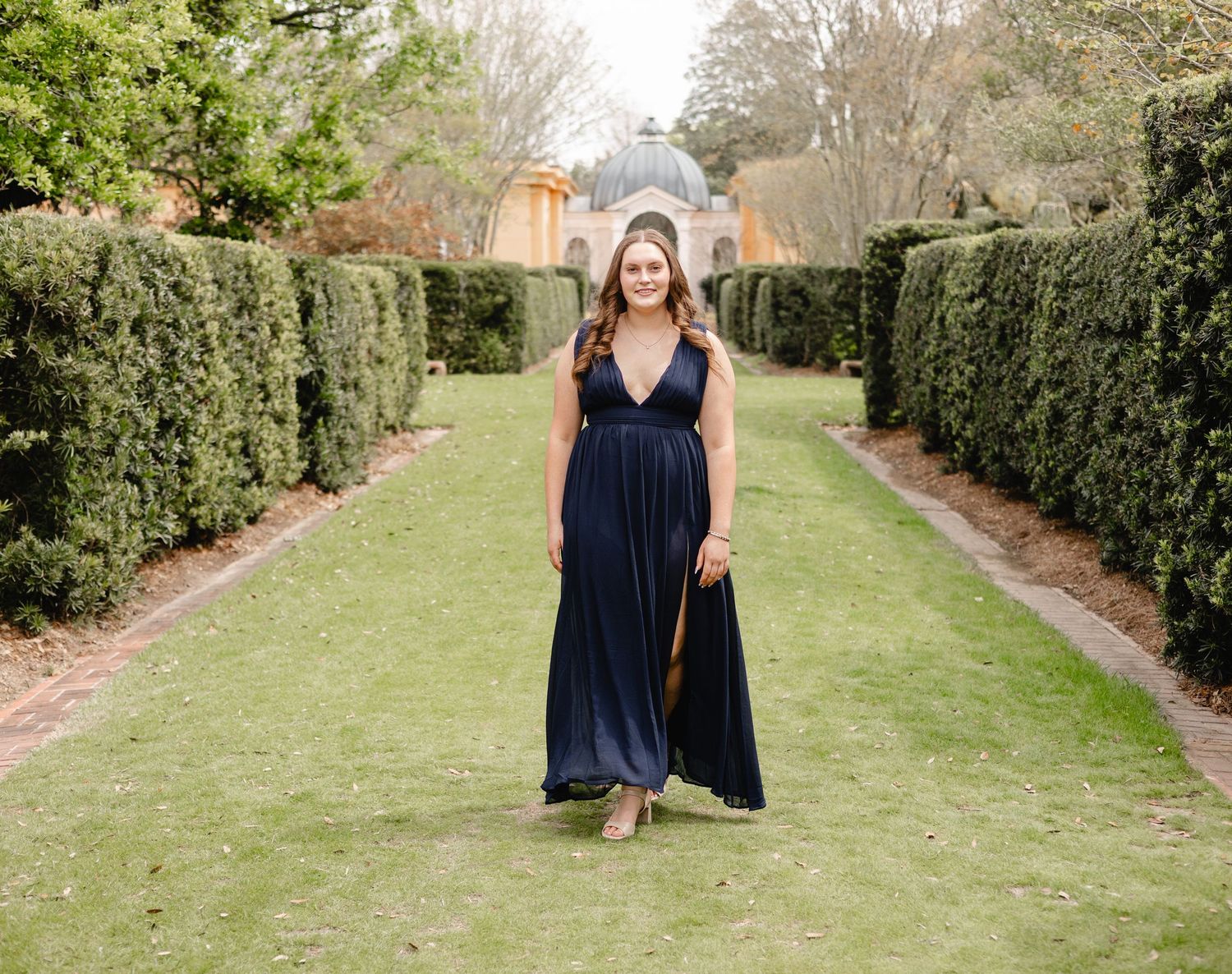 Woman in navy blue gown standing in formal garden pathway with manicured hedges and historic building.