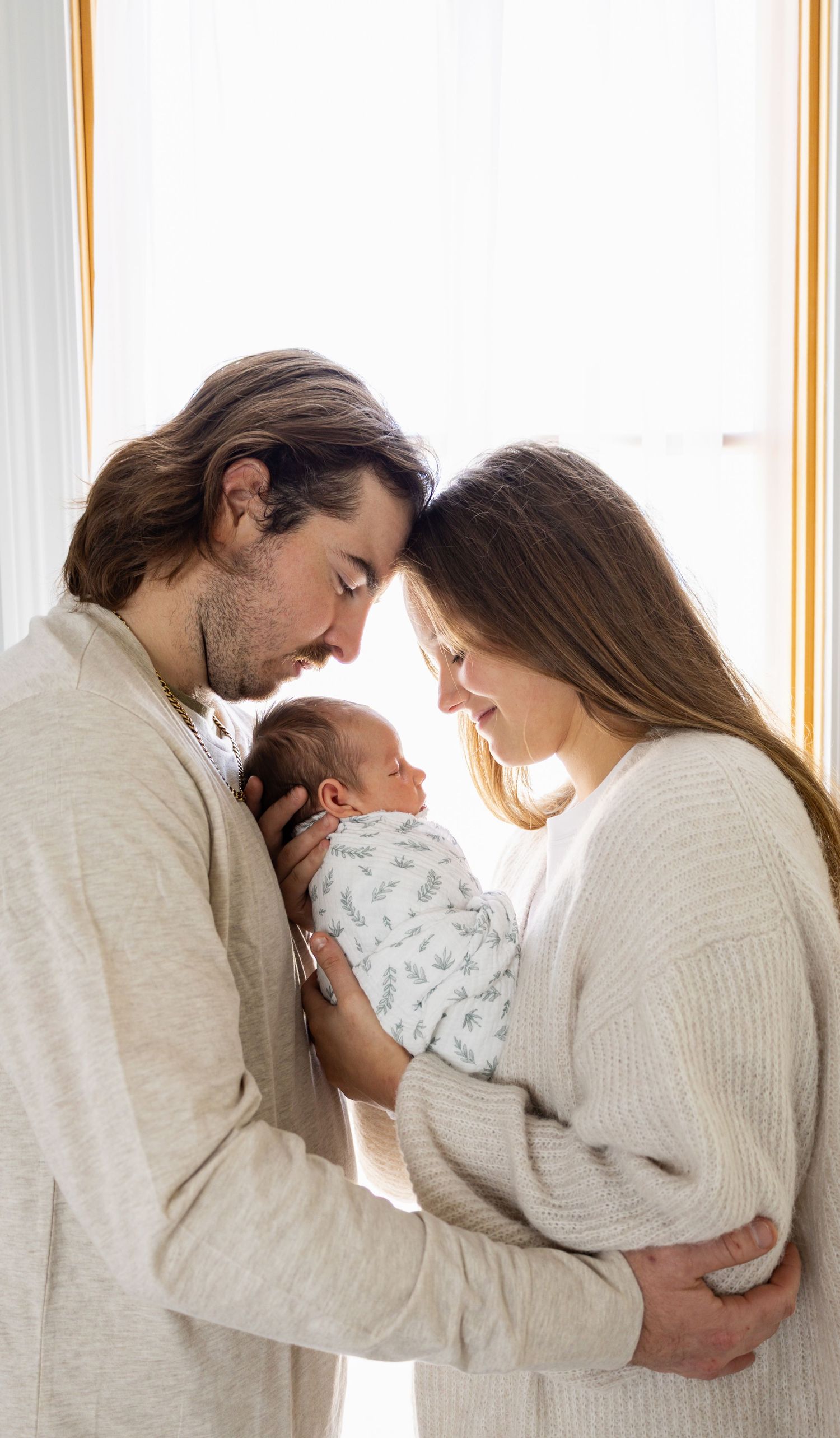 New parents tenderly hold their swaddled newborn baby together, touching foreheads in an intimate family portrait.