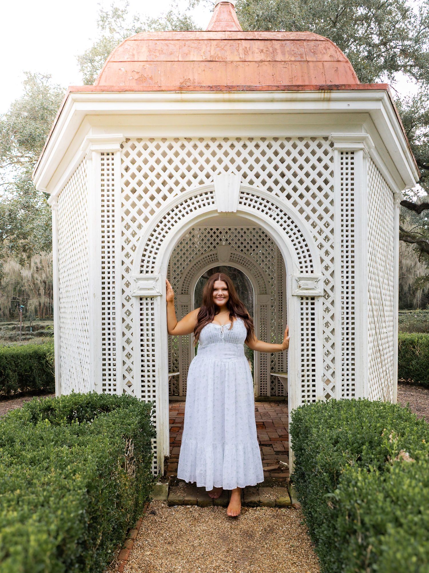 Woman in white midi dress posing in elegant white lattice garden gazebo with copper roof and manicured hedges.