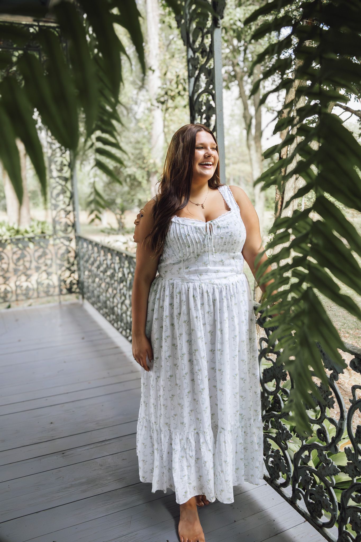 Woman in white floral maxi dress smiling on ornate iron porch surrounded by lush green ferns.