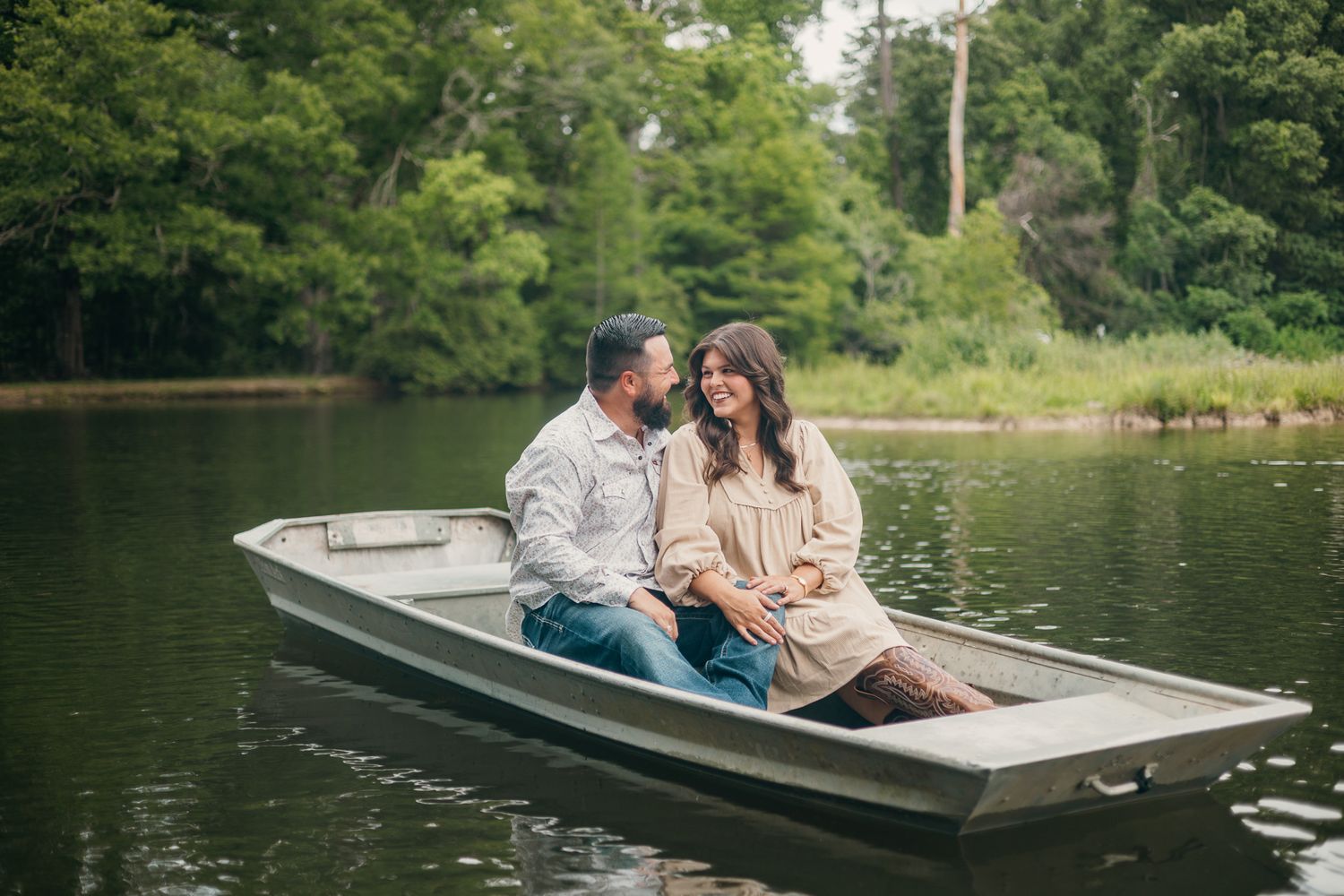 Couple sitting close together in a small aluminum rowboat on a calm lake surrounded by lush green trees.
