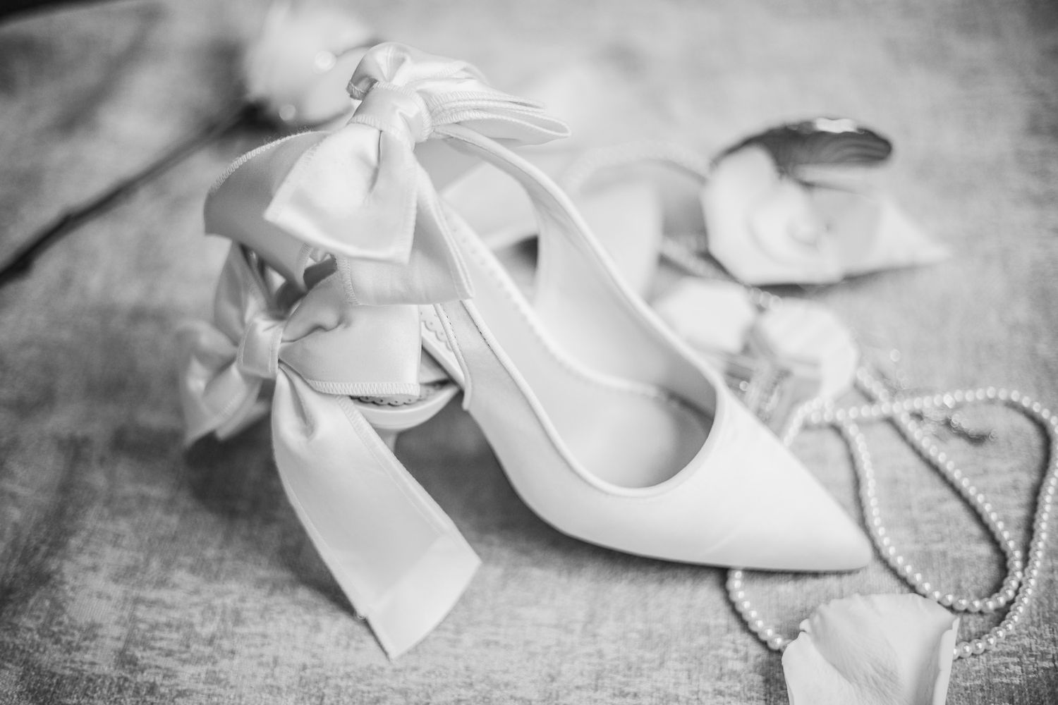 Black and white photo of elegant bridal heels with satin bow ribbons and pearl necklace on soft fabric surface.