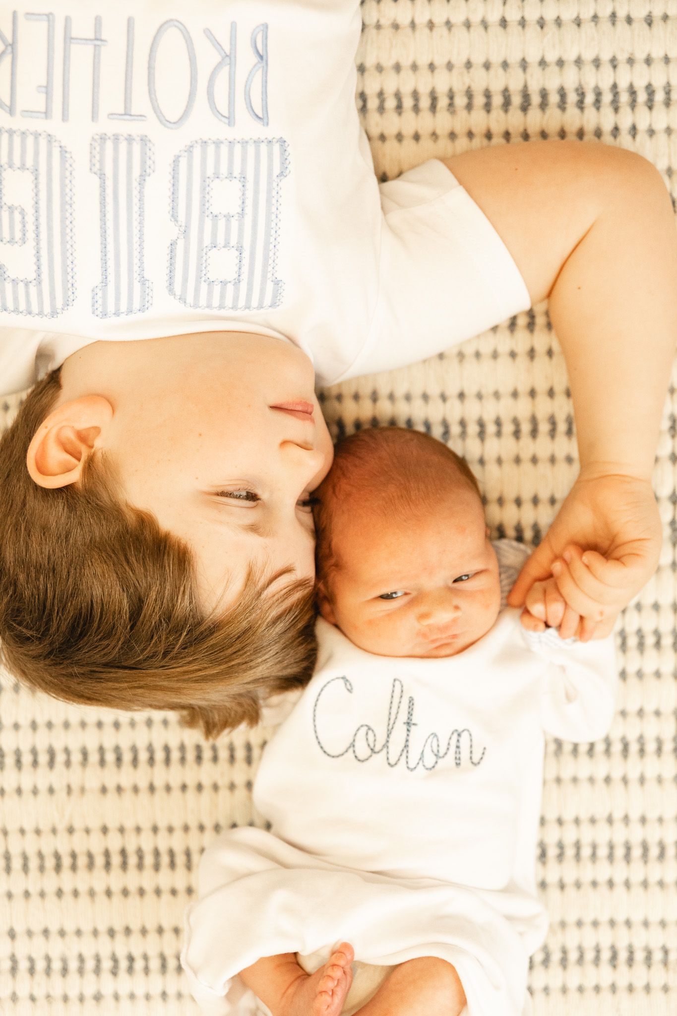 Older brother lying next to newborn baby Colton on a patterned blanket, both wearing matching white outfits.