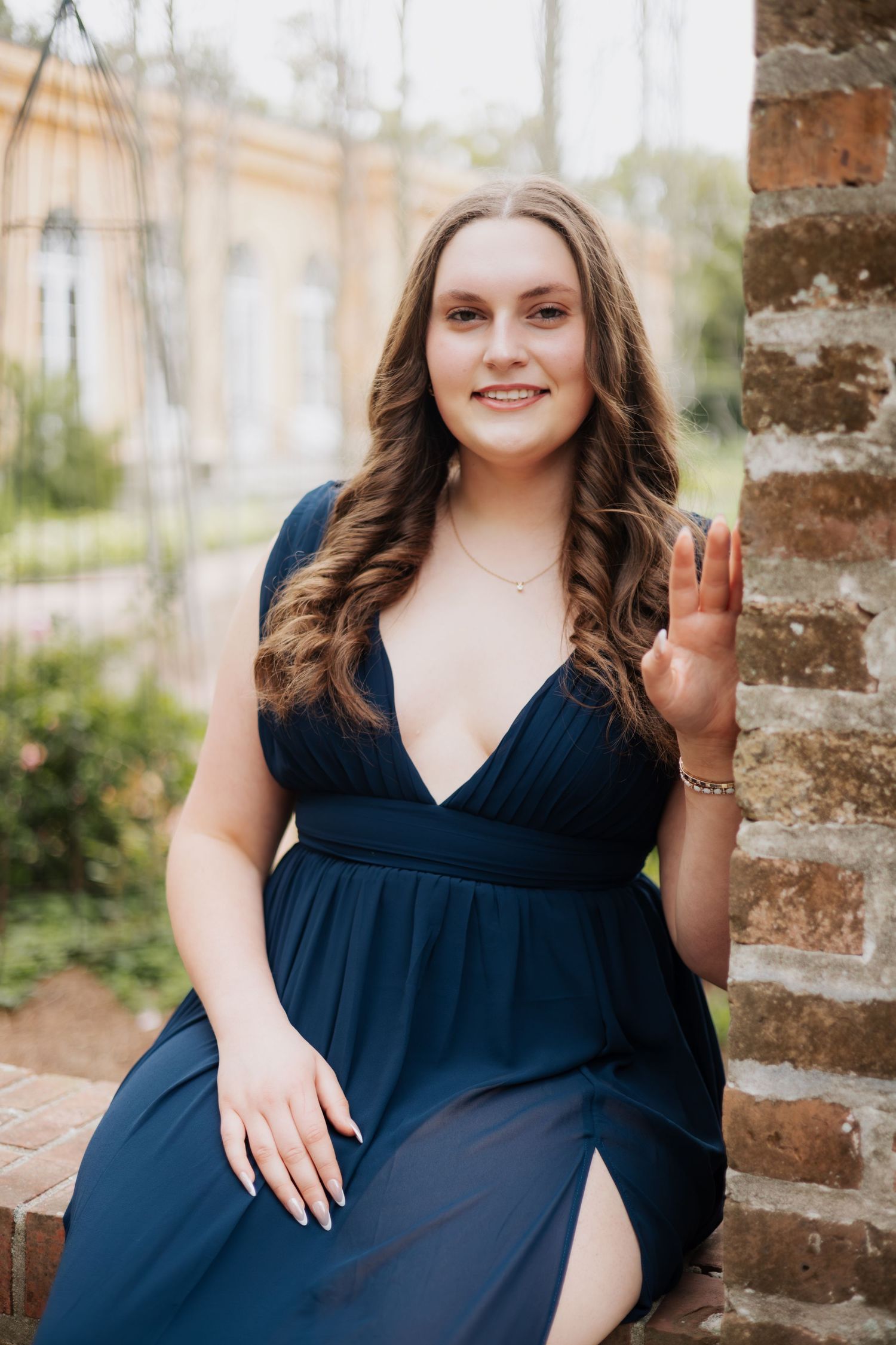 Young woman in navy blue dress waving and smiling while seated beside a brick column outdoors.