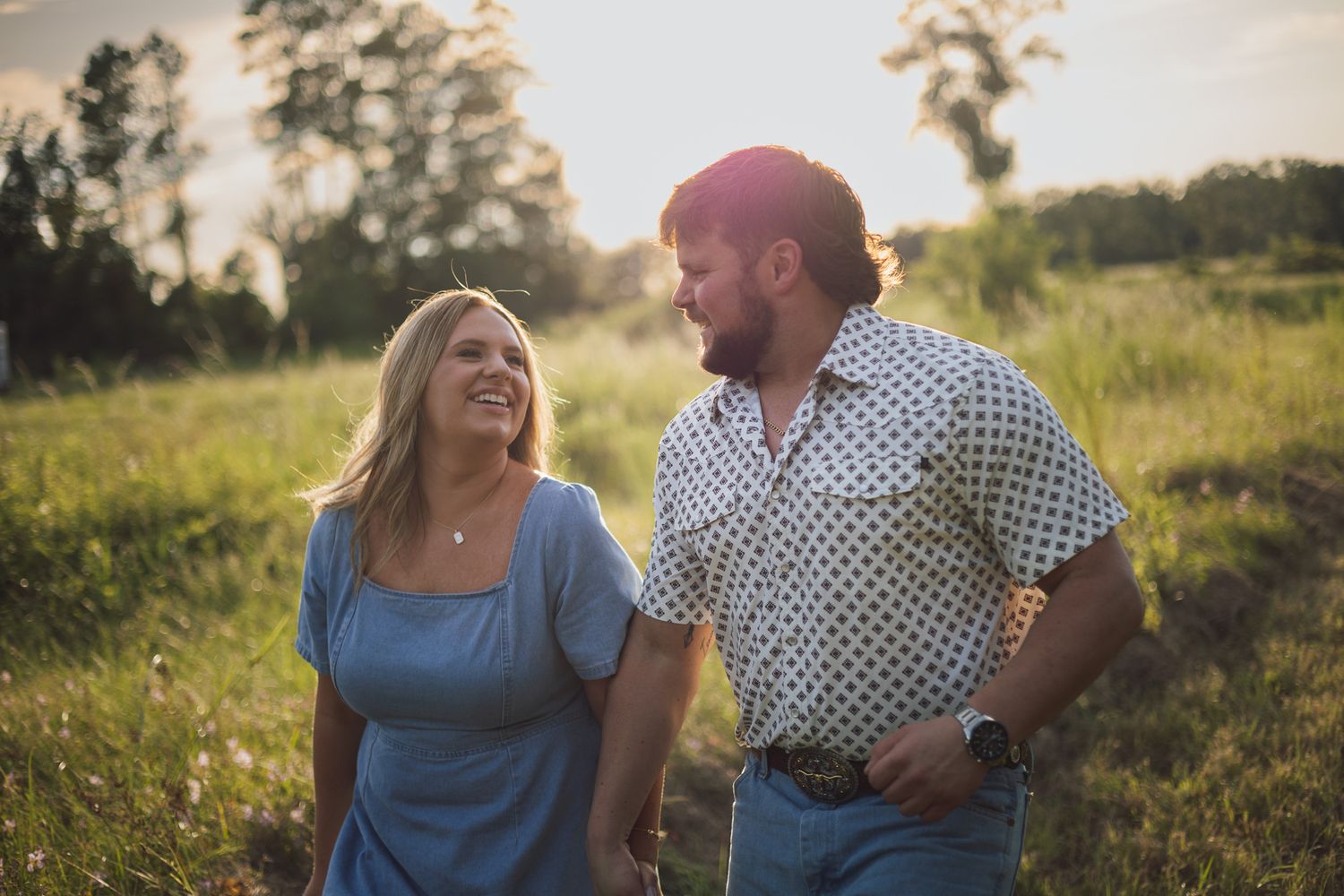 Couple walking hand in hand through a sunny green field, smiling at each other in golden hour light.