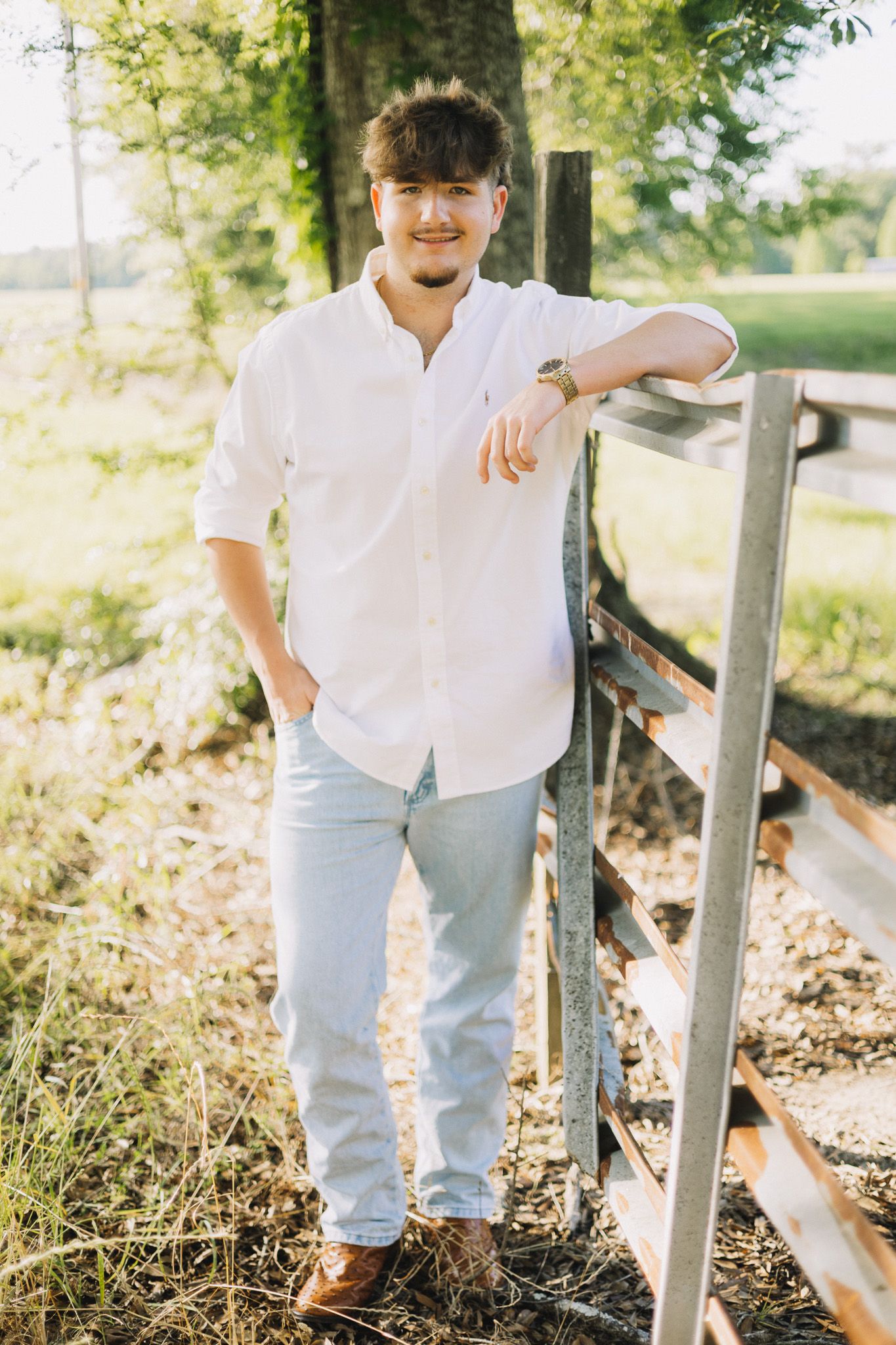 Young man in white shirt and light jeans leaning on a wooden fence outdoors in a sunny green setting.