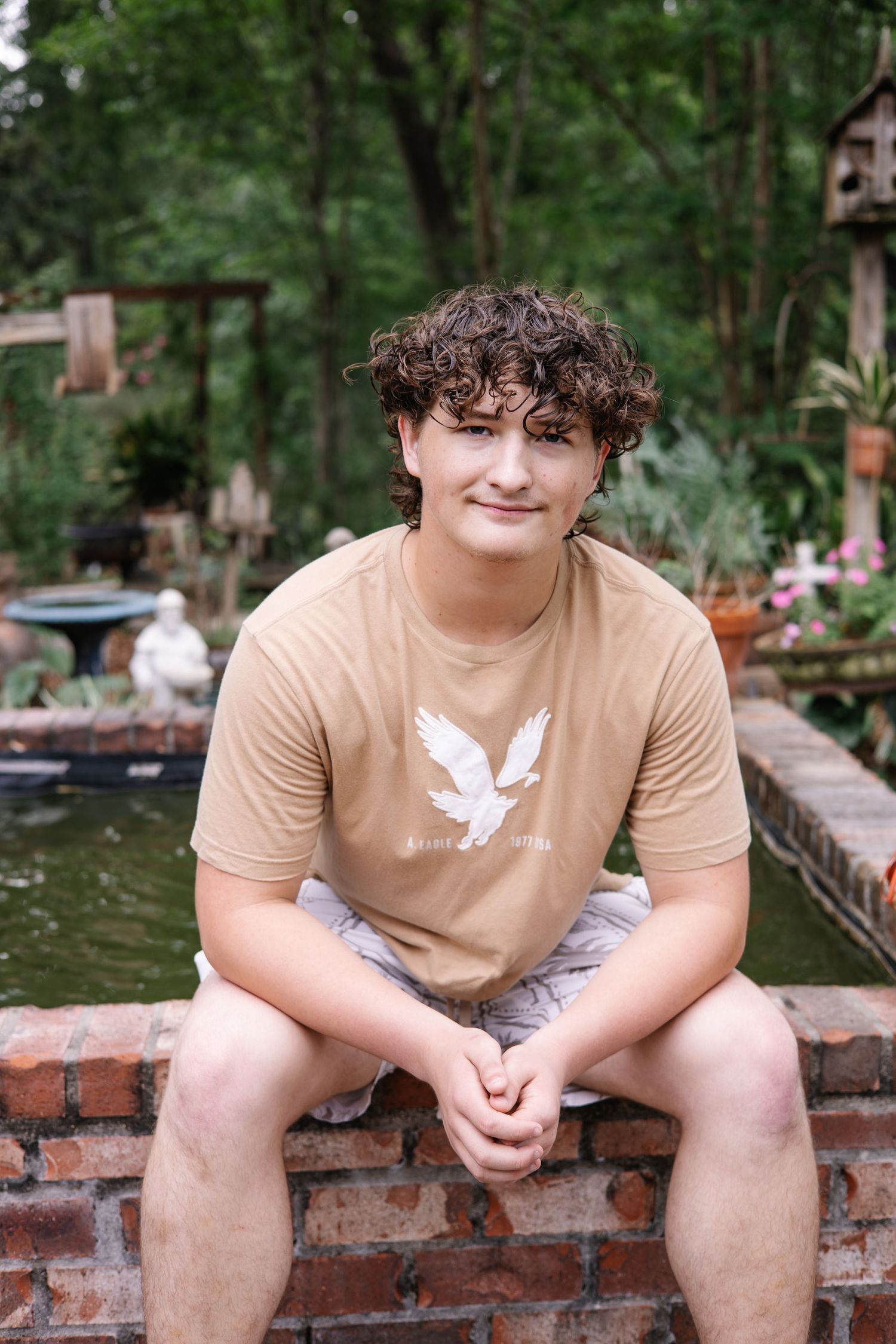 A smiling teenage boy with curly hair sits on a brick wall in a lush garden, wearing a tan American Eagle t-shirt.