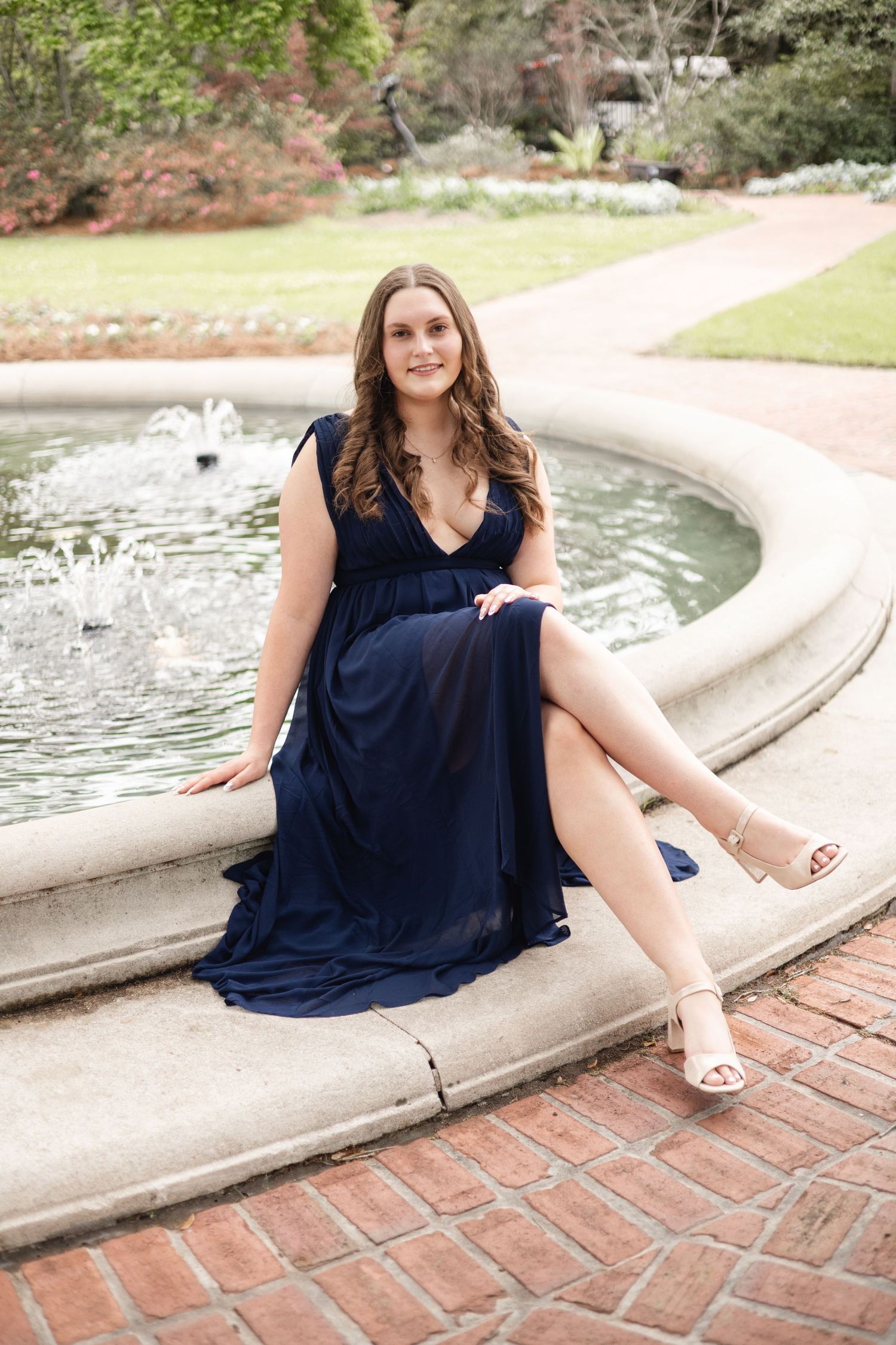 Young woman in navy blue maxi dress seated on stone fountain edge in garden setting.