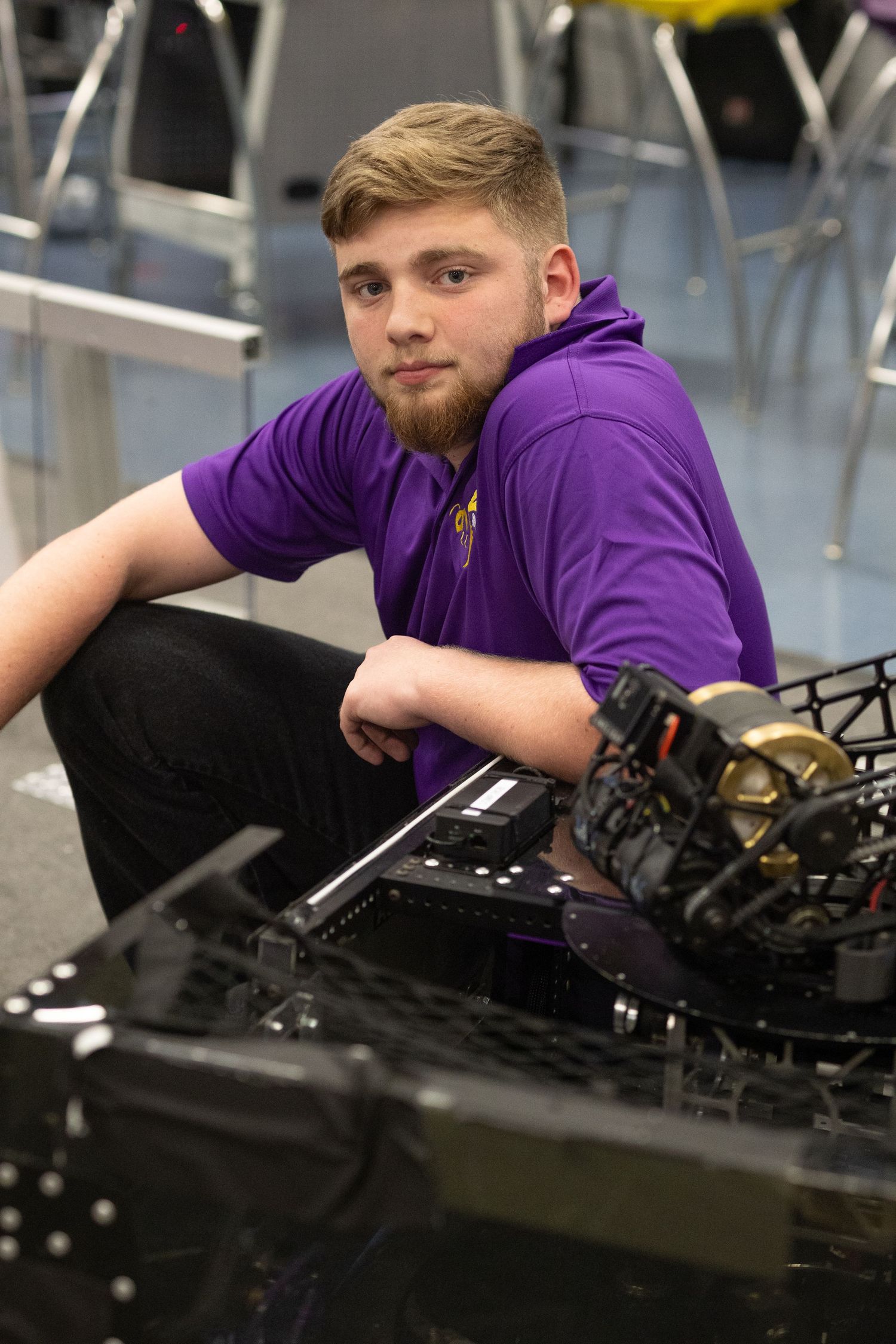 Young man in purple polo shirt sitting on floor next to robotics equipment in a workshop setting.