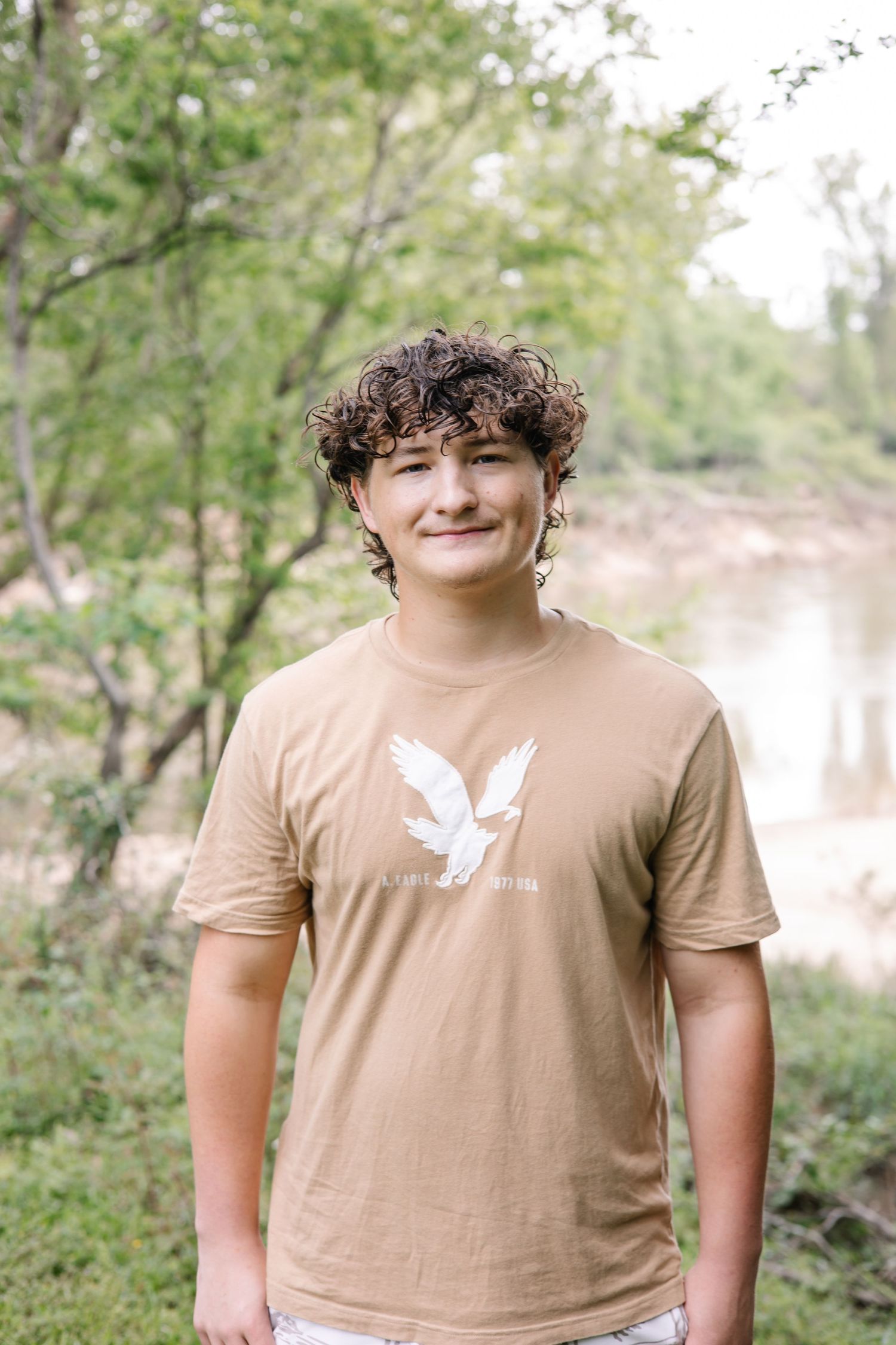 Smiling teen with curly hair wearing a tan American Eagle t-shirt, standing outdoors near trees and a river.
