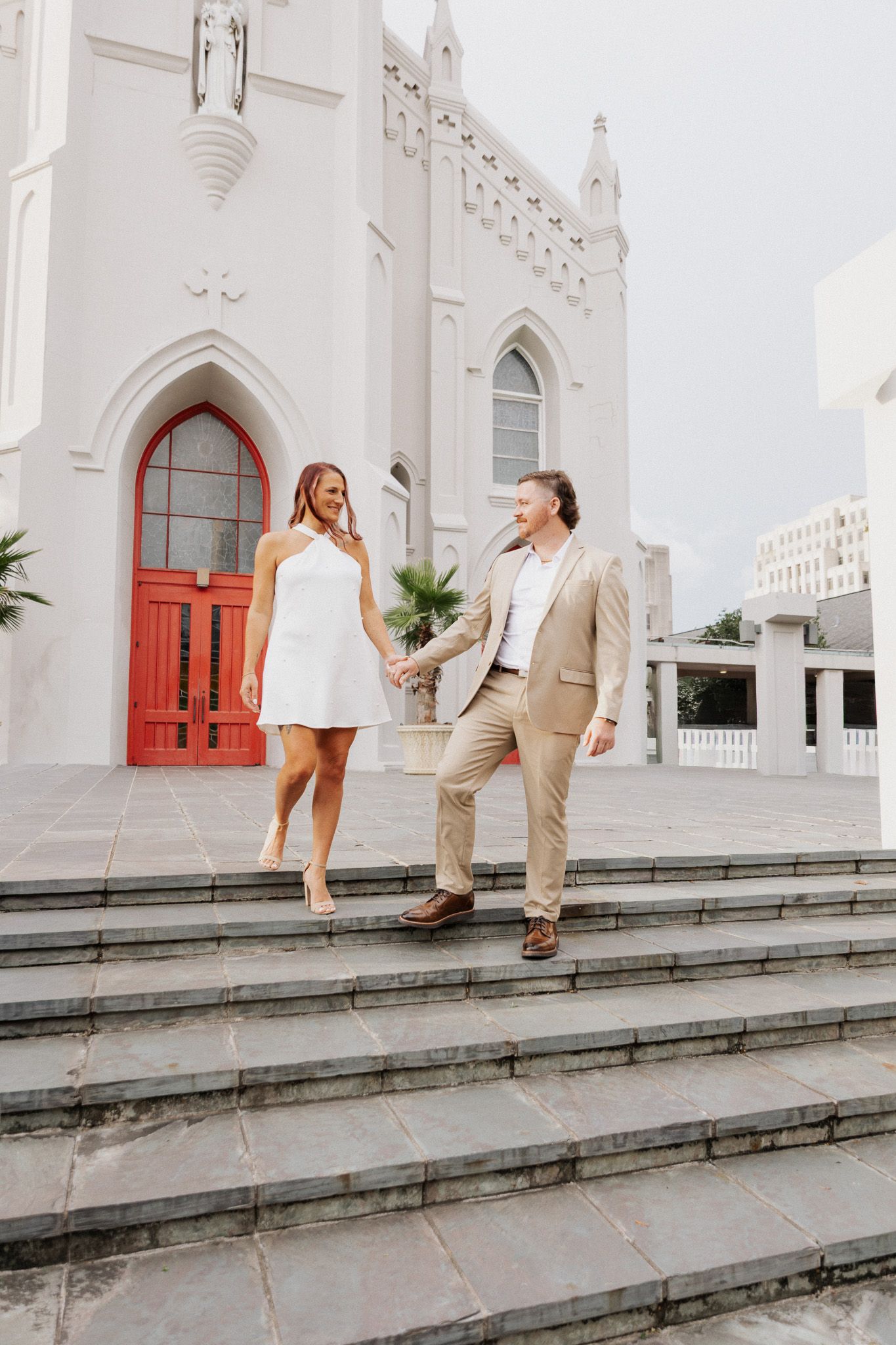 Couple holding hands on church steps, woman in white dress, man in tan suit, white Gothic church backdrop.
