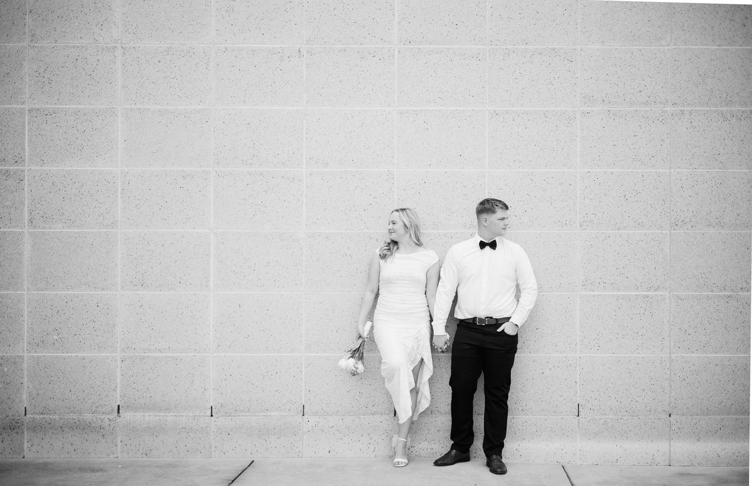 Black and white photo of a bride and groom holding hands against a stone wall in elegant wedding attire.