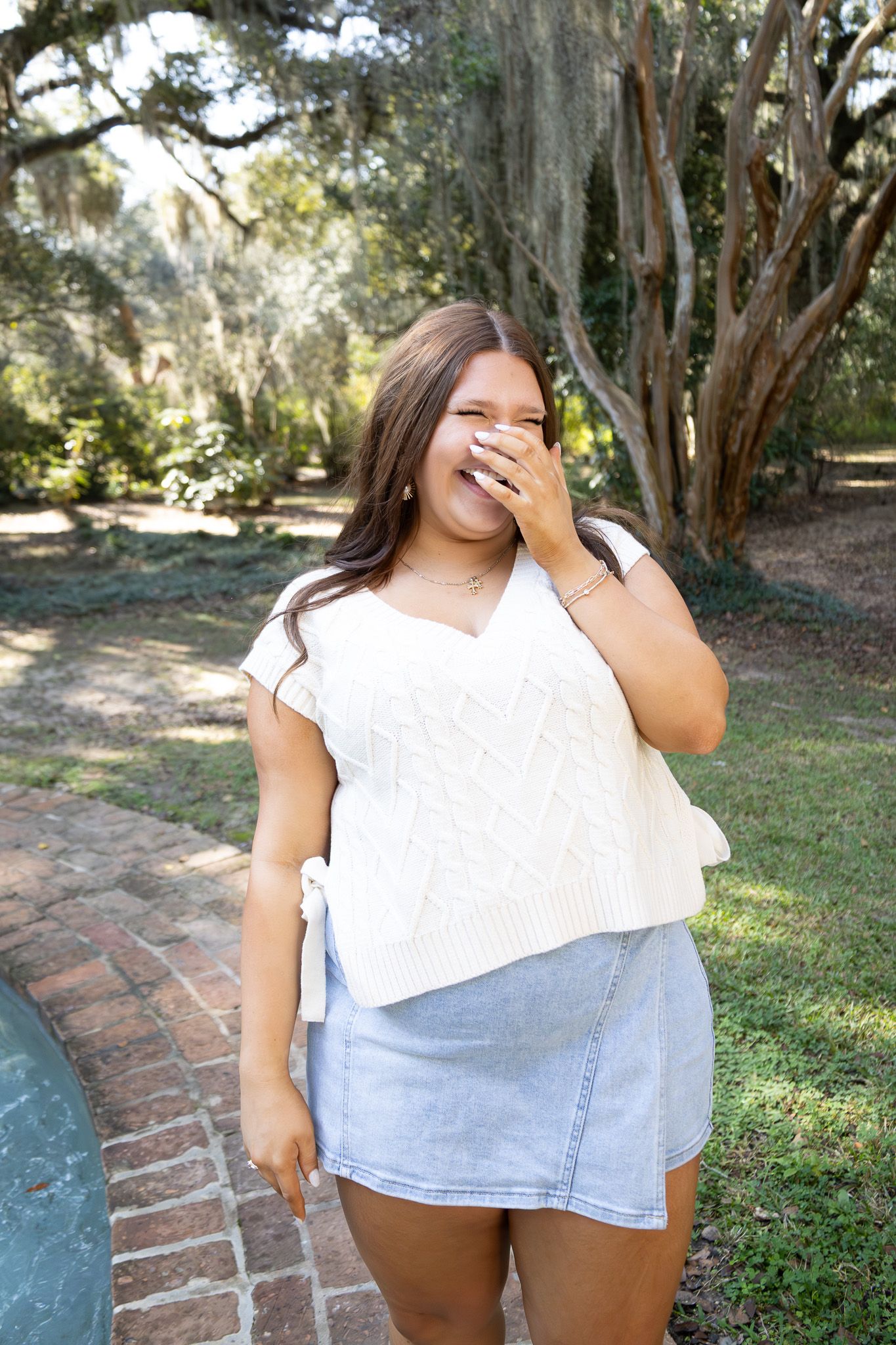 Young woman in white knit top and denim skirt laughing outdoors in a lush green park setting.