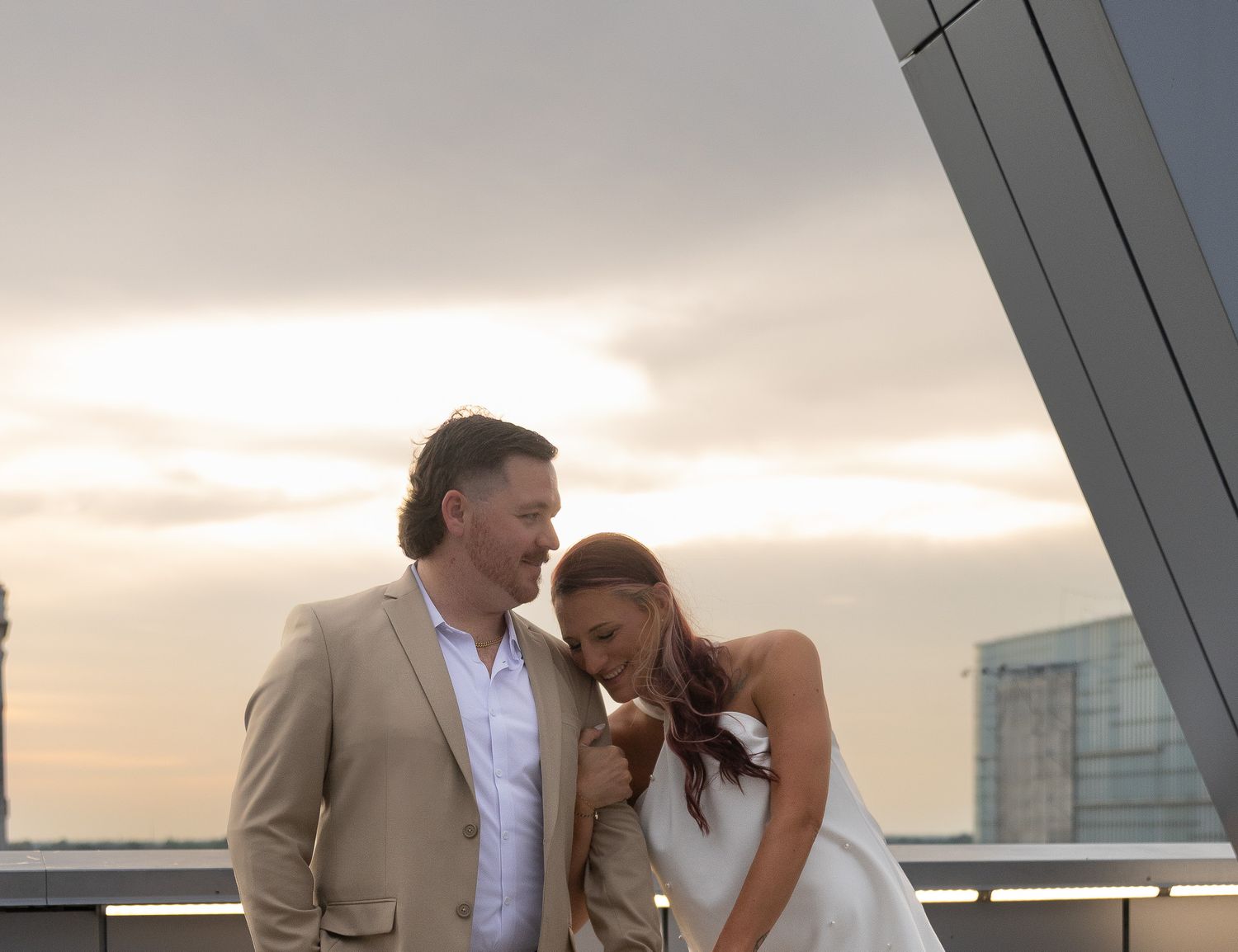 Couple in wedding attire sharing a romantic moment at sunset on a rooftop, man in beige suit, woman in white dress.
