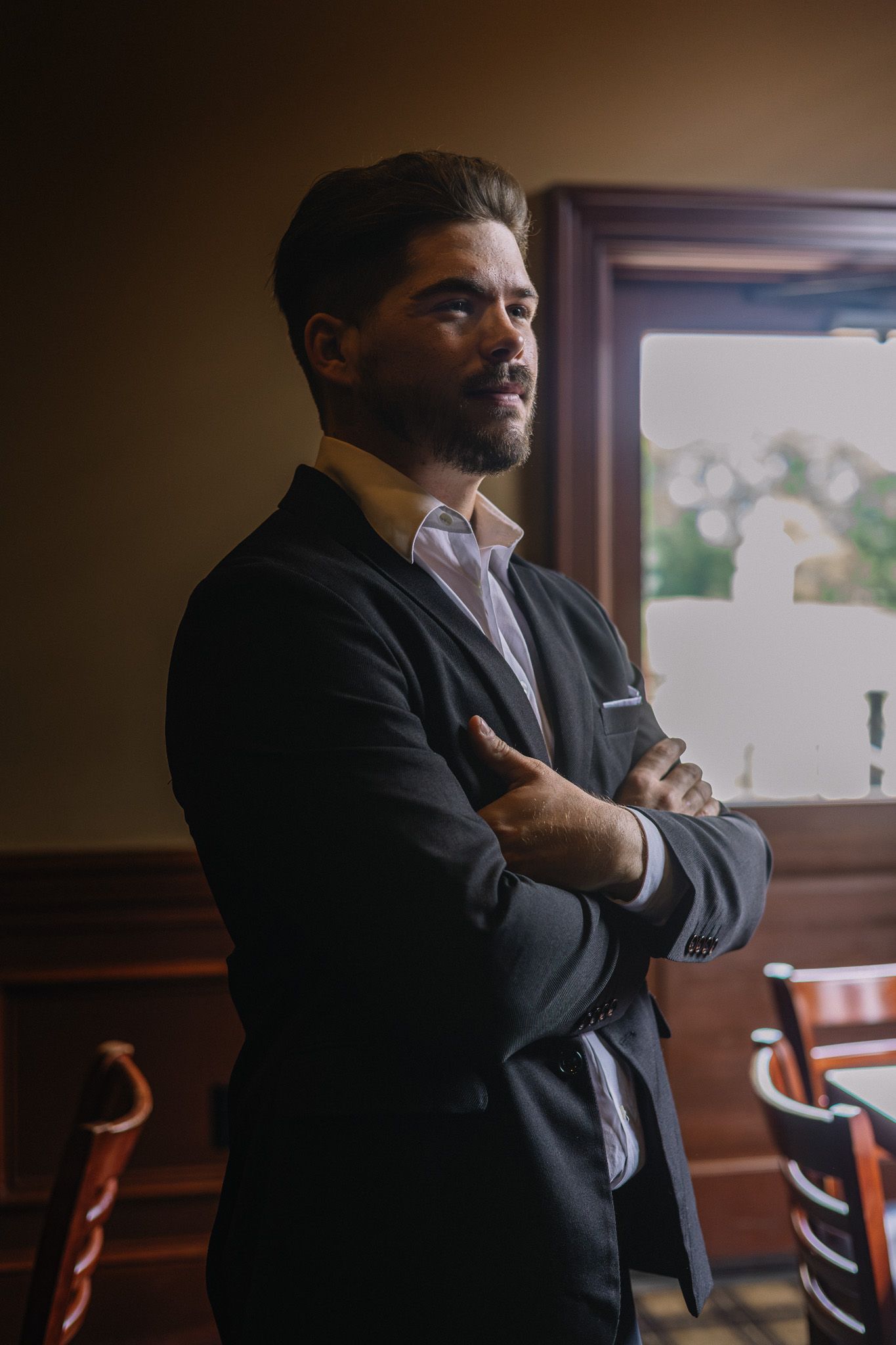 A confident bearded man in a dark suit stands with arms crossed near a window in an elegant interior setting.