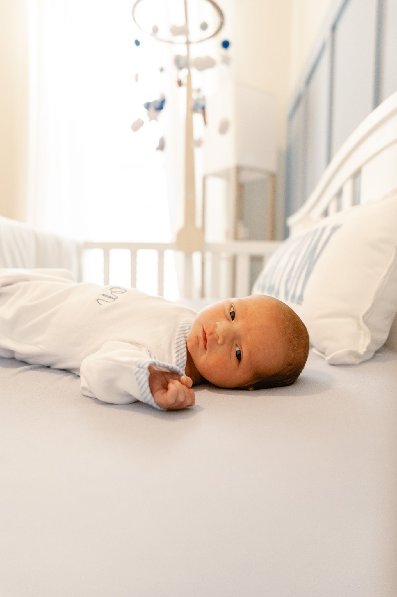 Newborn baby in white onesie lying awake on white crib mattress with mobile hanging above in bright nursery.