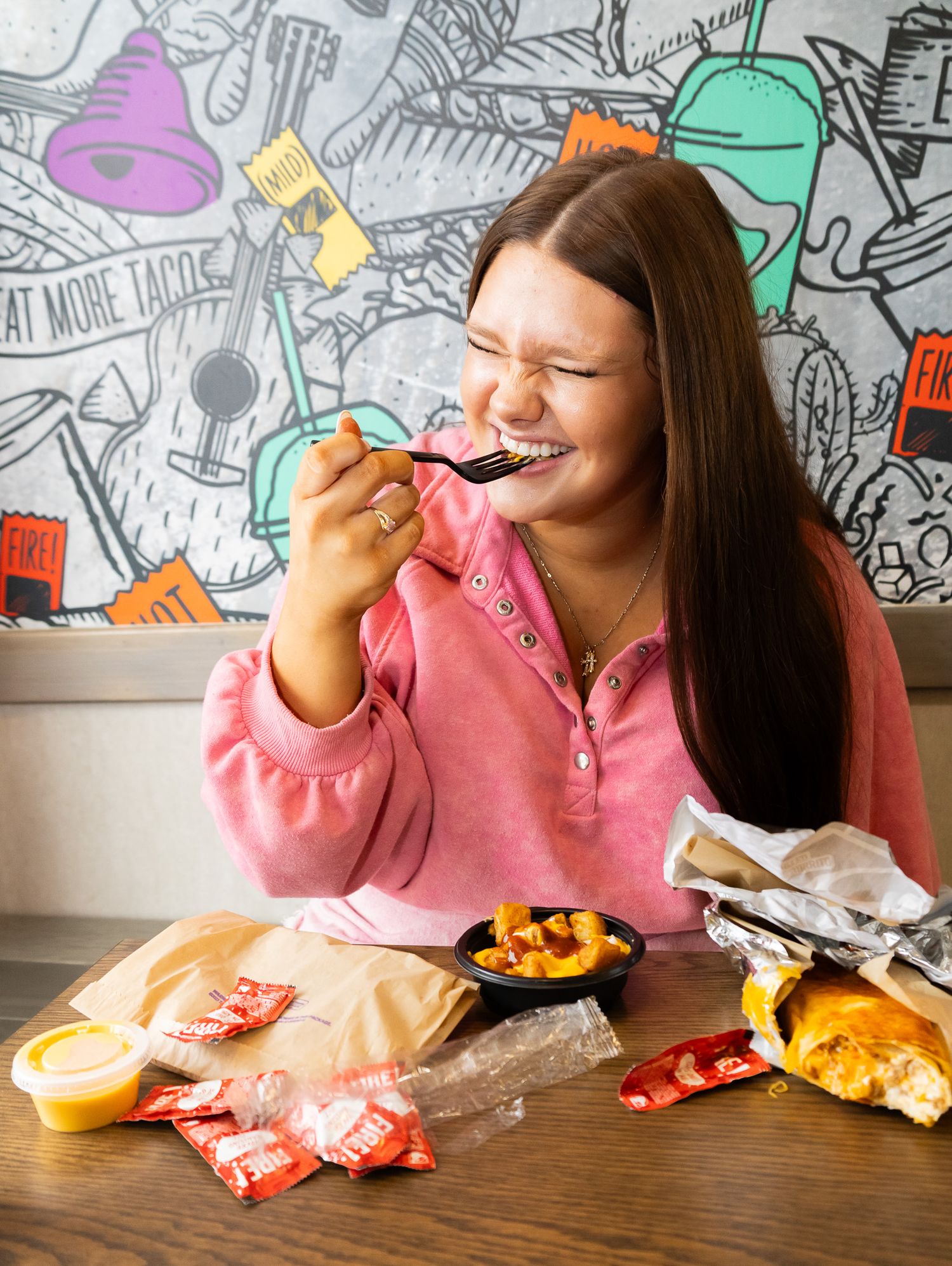 Woman in pink hoodie smiling while eating at Taco Bell, enjoying nachos with sauce packets on table.
