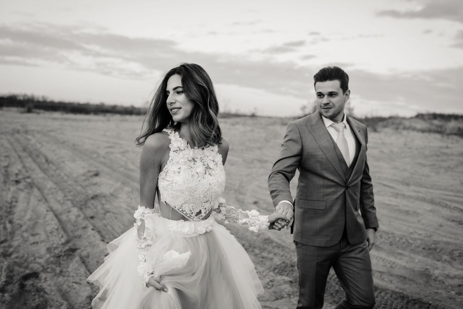 Candid black and white photo of couple walking through empty field in wedding attire.