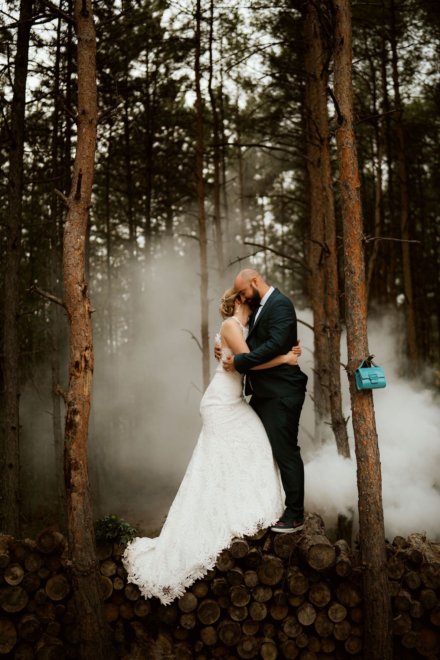 Romantic wedding portrait in misty forest with white smoke and stacked logs creating dreamy woodland atmosphere.