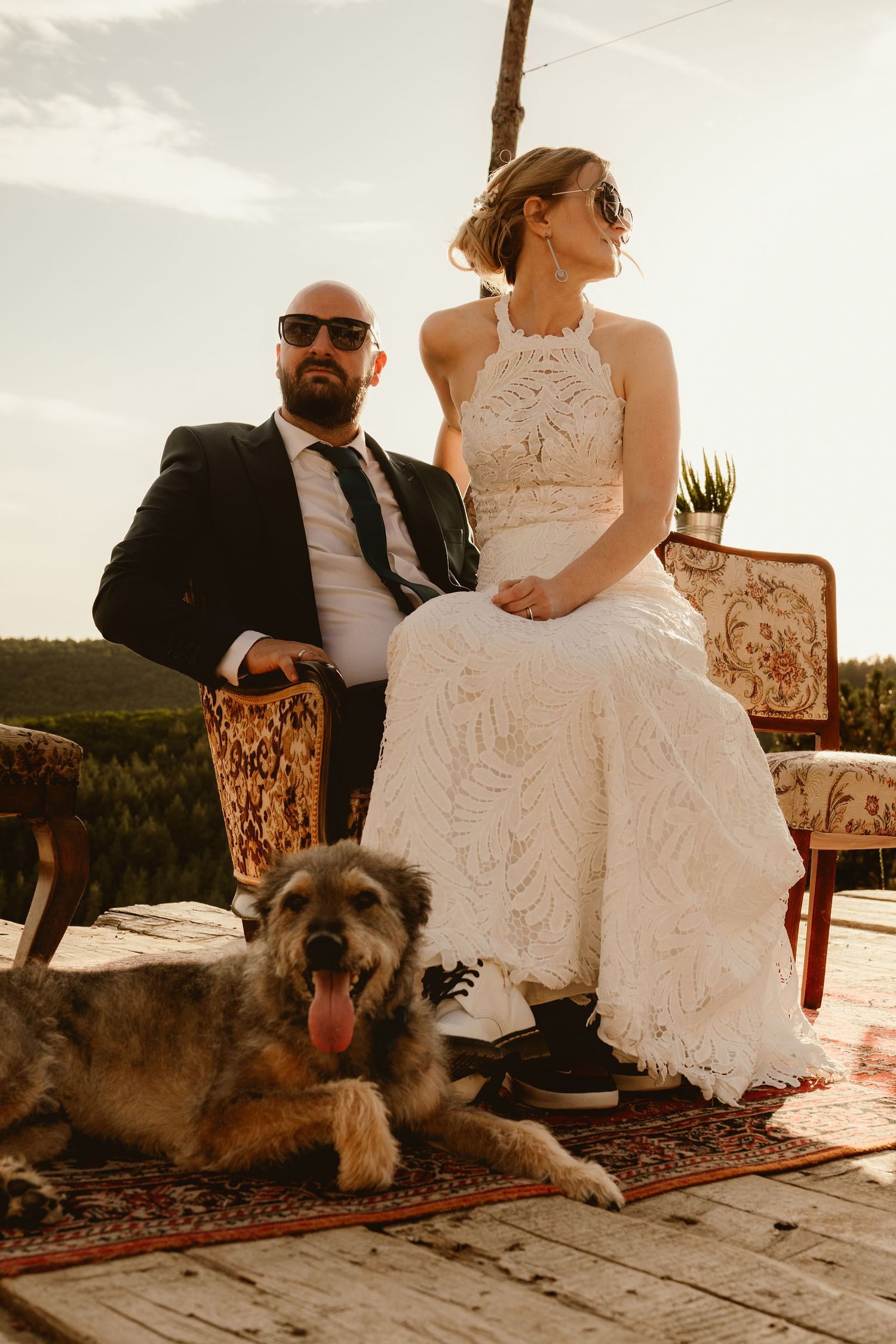 A couple sits on vintage chairs on an outdoor deck with their happy dog resting at their feet on an ornate rug at sunset.