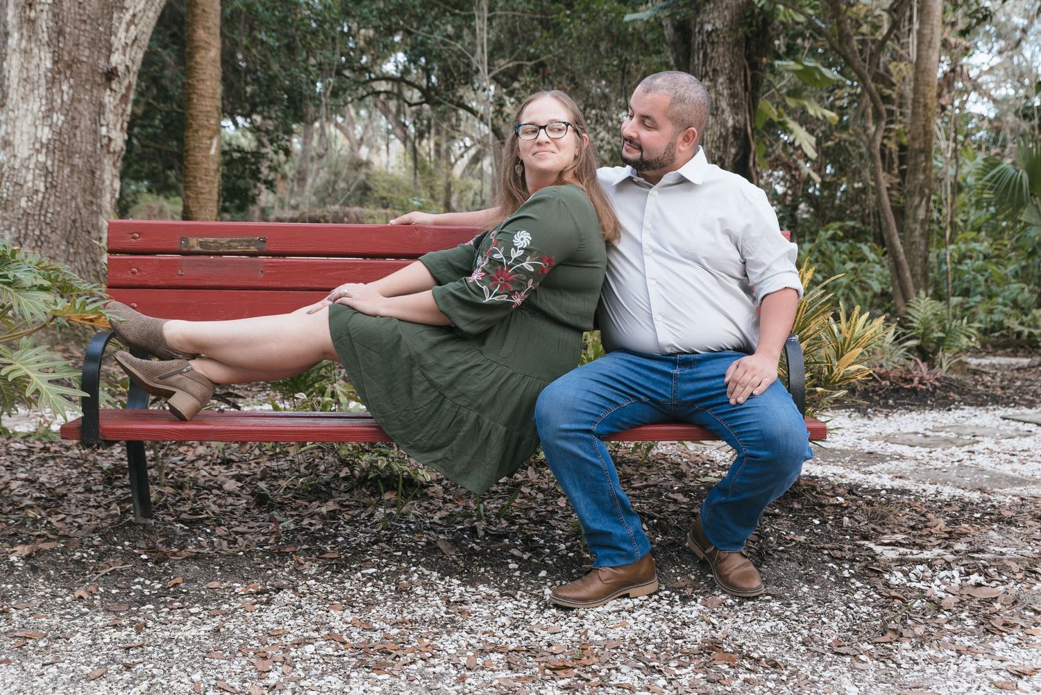 Couple sitting on red bench in wooded park setting during photo session.