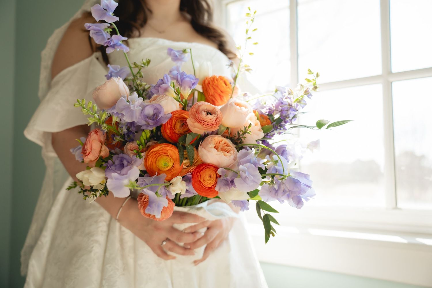 Bride holding a vibrant bouquet of orange ranunculus and purple wildflowers in a bright white room.