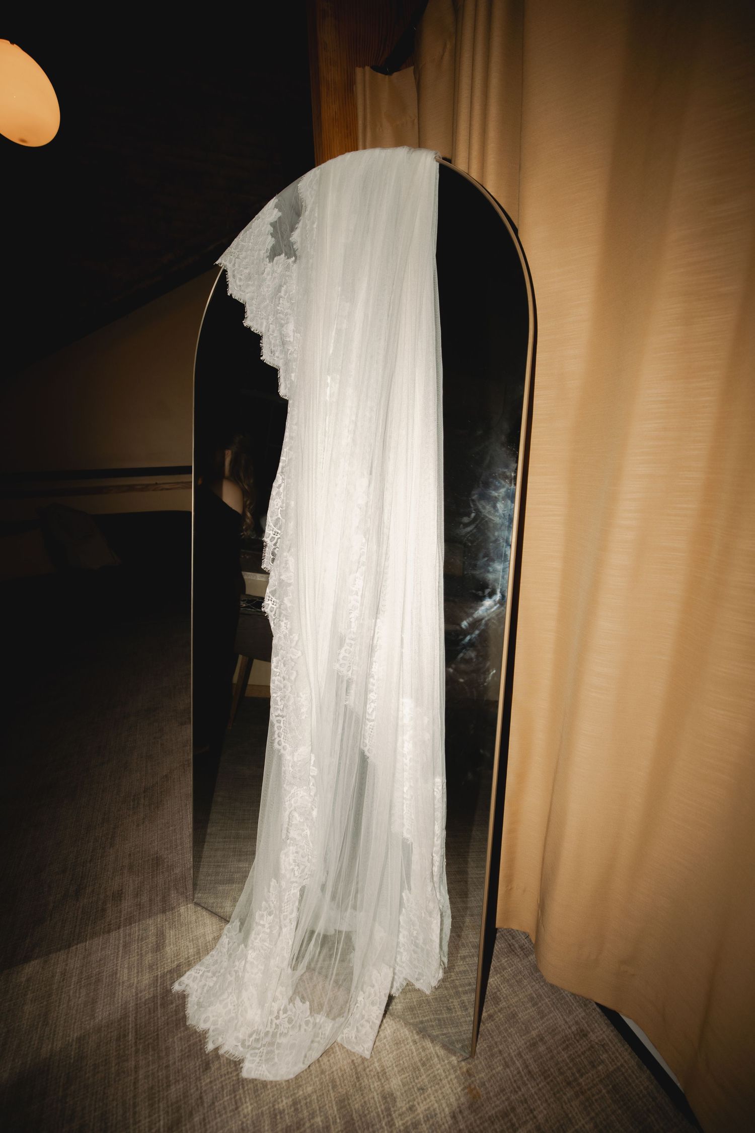 Elegant white lace wedding veil draped over an arched mirror in a dimly lit room.