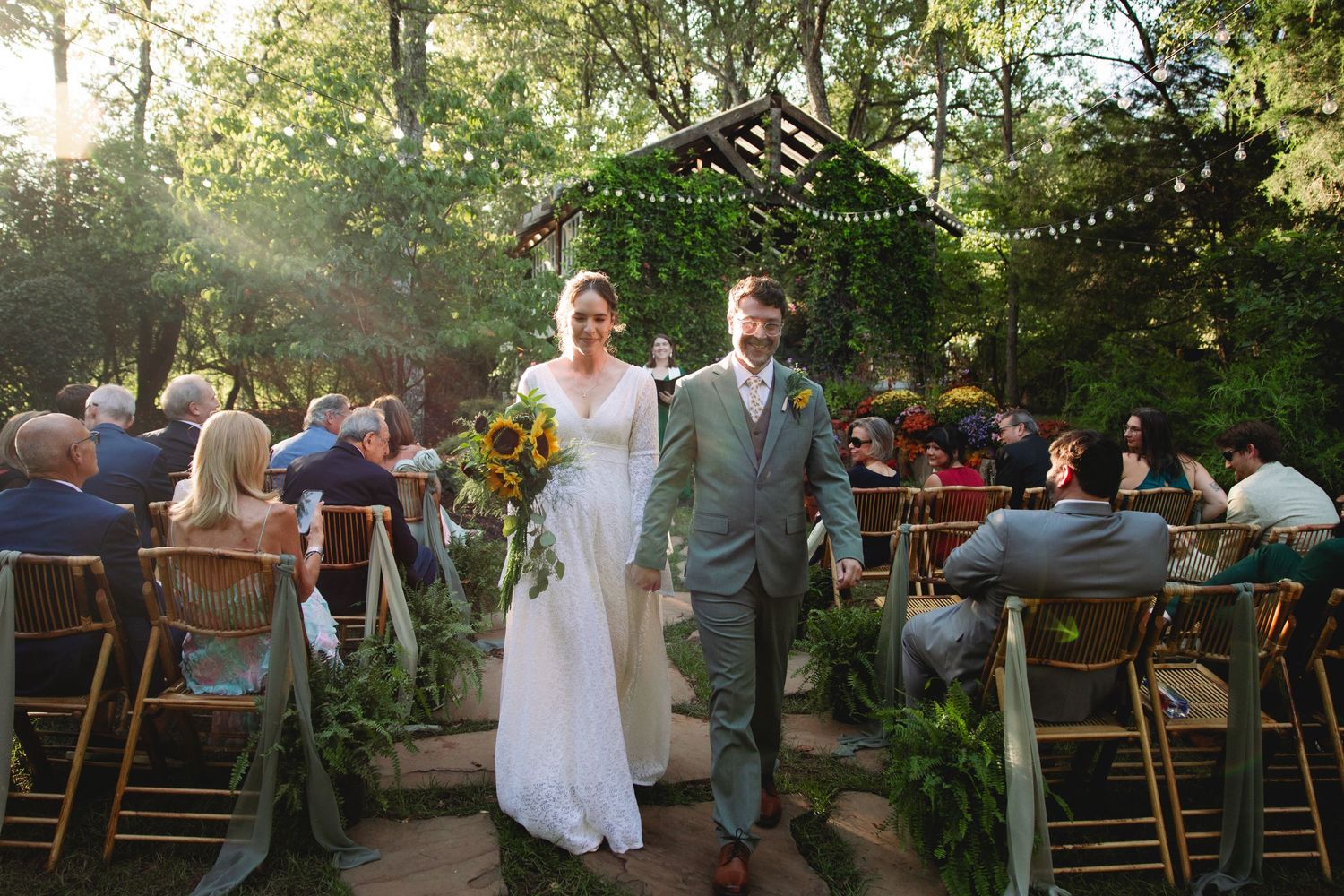 Bride and groom walk down outdoor wedding aisle surrounded by lush greenery and seated guests in a sunlit garden.