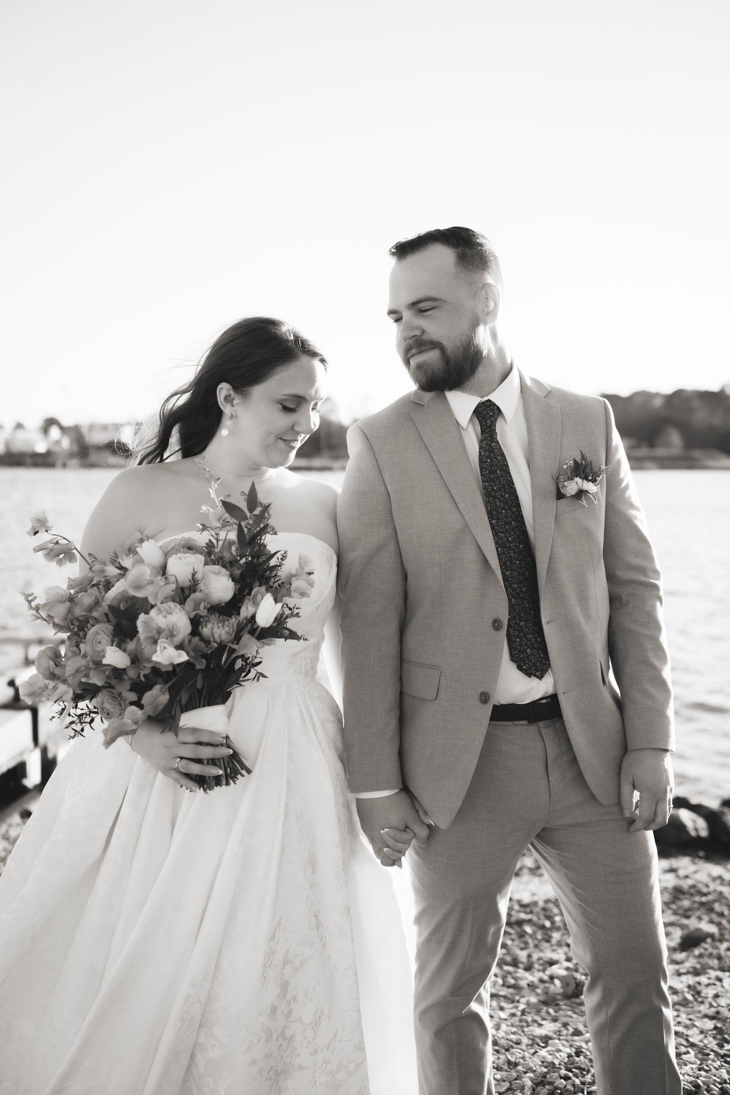 Black and white photo of bride and groom holding hands near waterfront on wedding day.