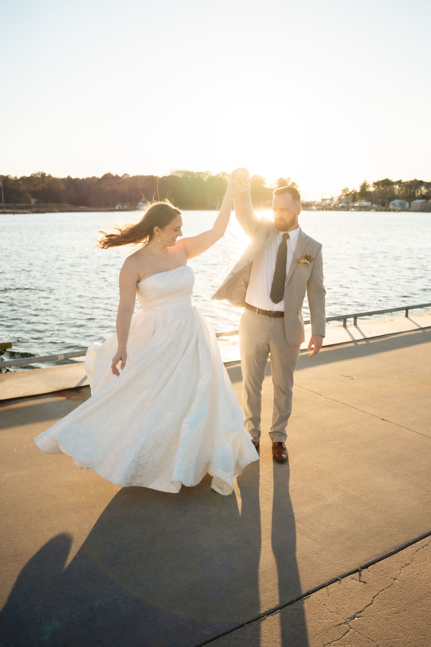 Bride and groom dance joyfully on waterfront boardwalk bathed in golden sunset light.