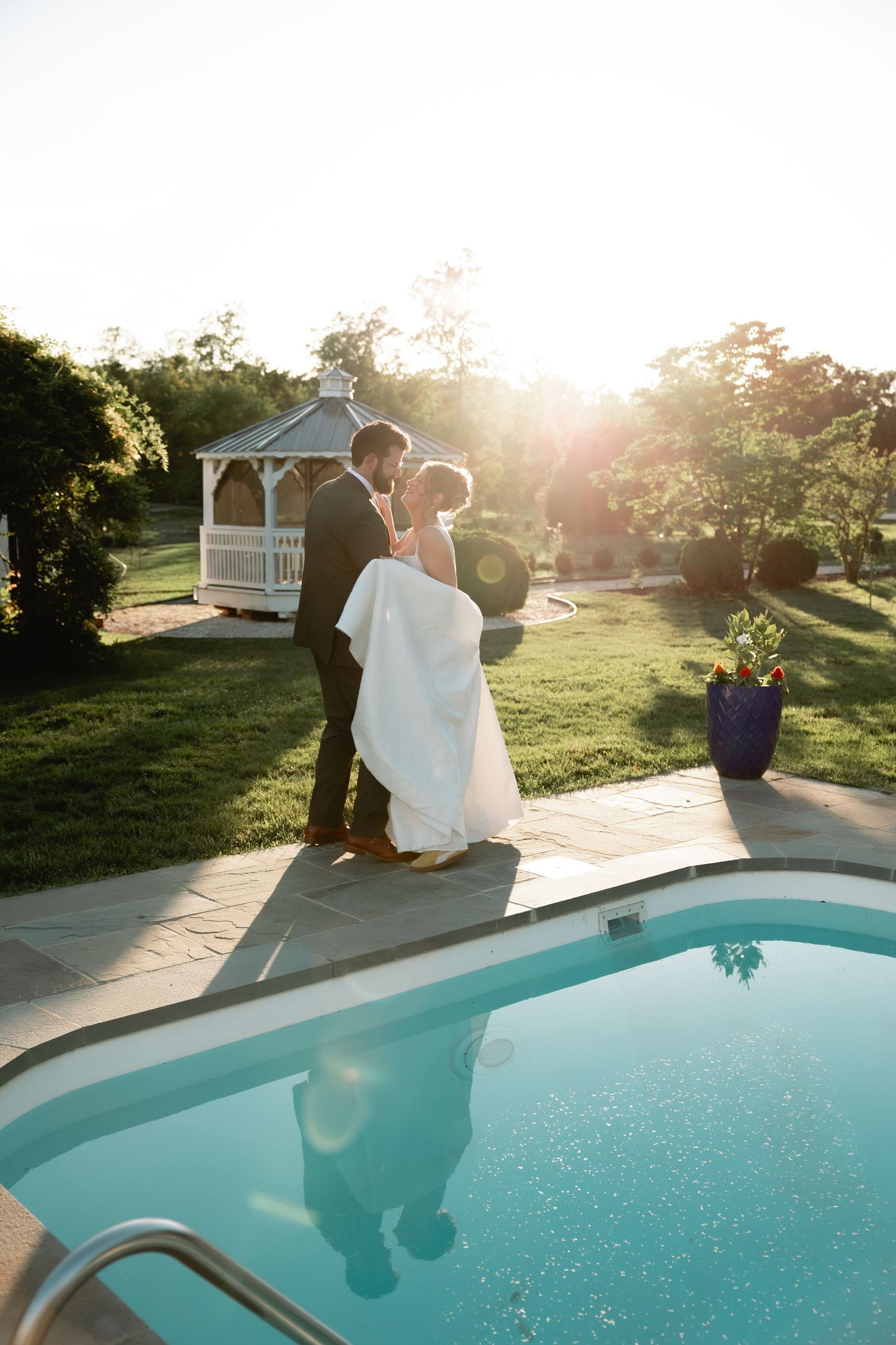 Bride and groom share a romantic moment by a pool at golden hour, with a white gazebo in the background.