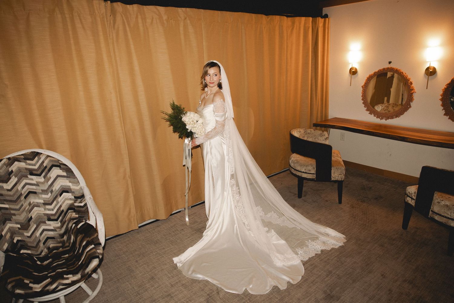 Bride in a long white wedding gown and cathedral veil holding a bouquet in an elegant dressing room.