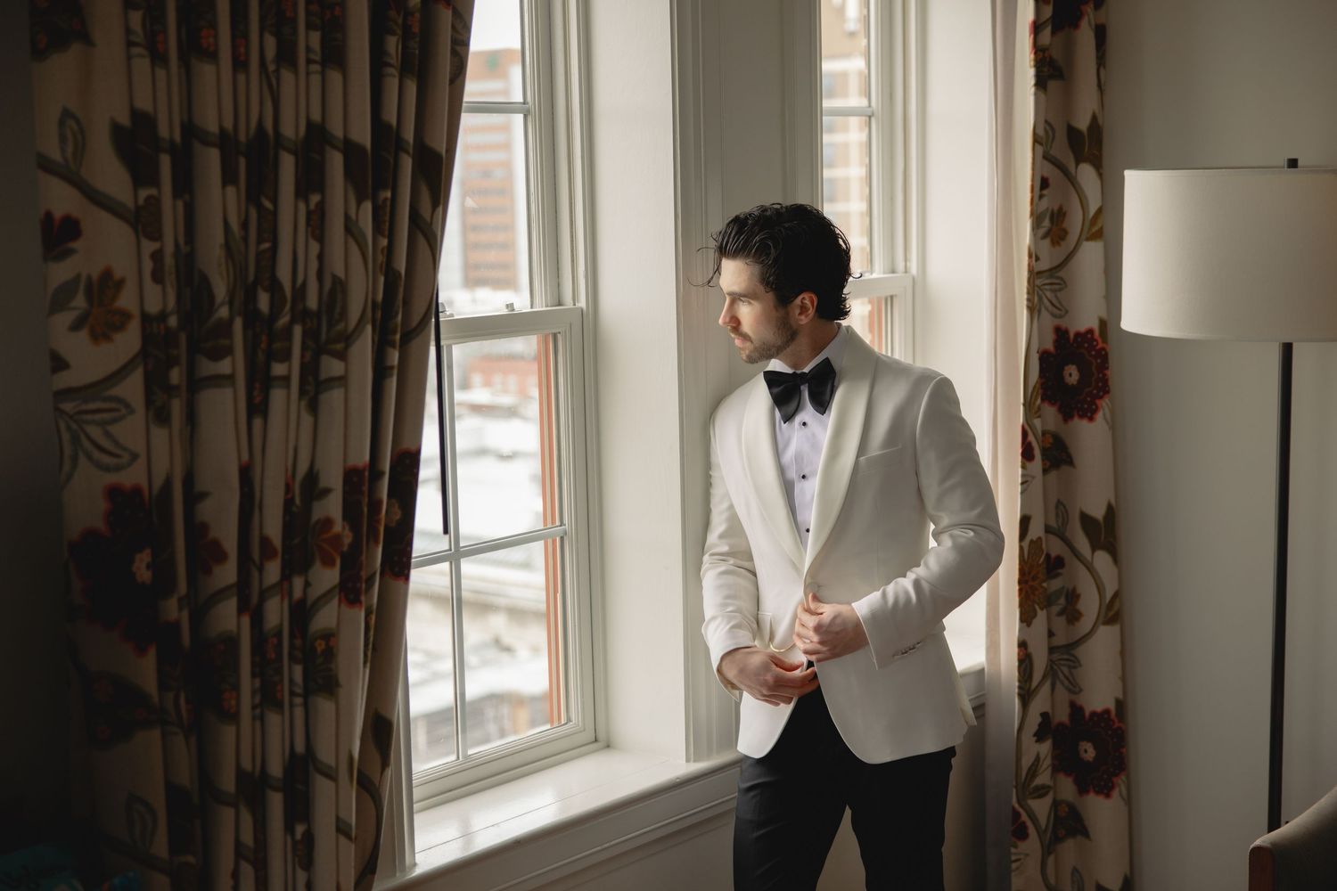 Dapper groom in a white tuxedo jacket gazing thoughtfully out a window with floral curtains.