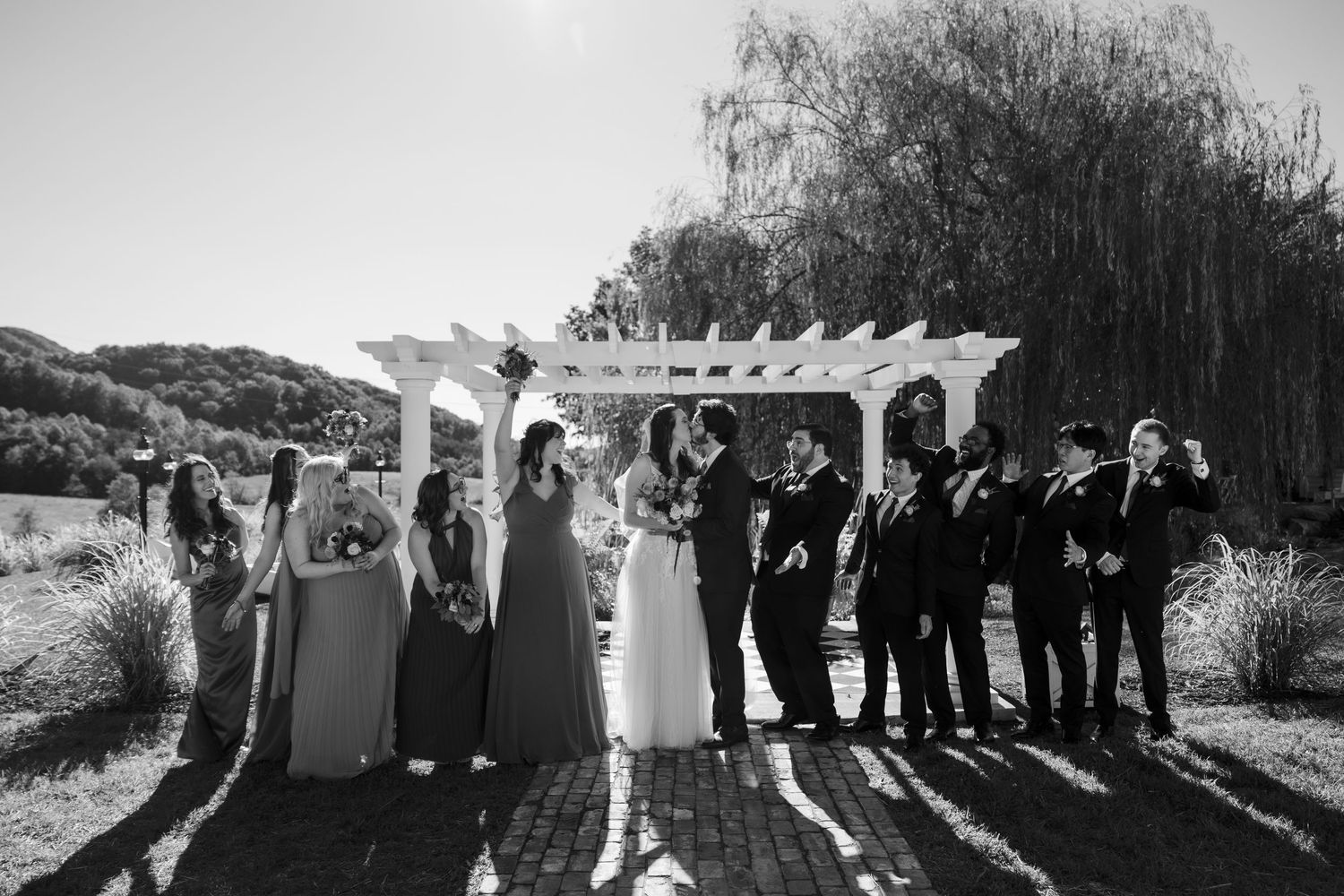 Black and white wedding party photo under pergola with bride and groom kissing flanked by bridesmaids and groomsmen.