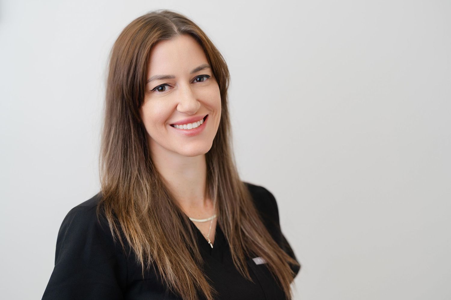 Professional woman with long brown hair smiling, wearing a black top against a light background.