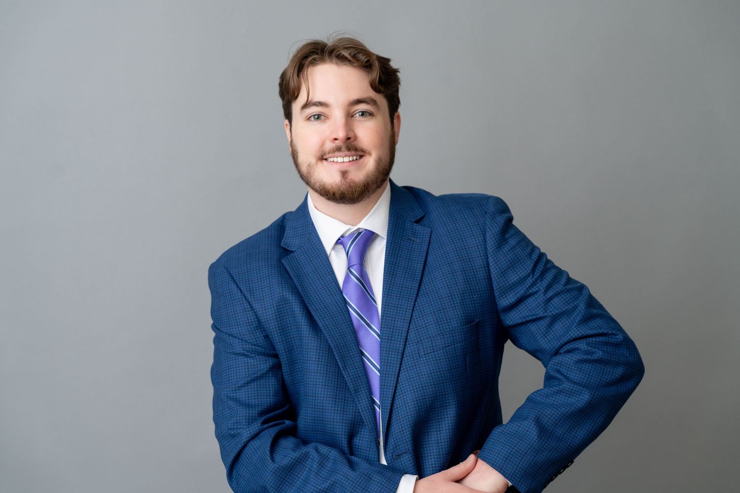 Professional man in blue suit and purple tie smiling confidently against gray background.