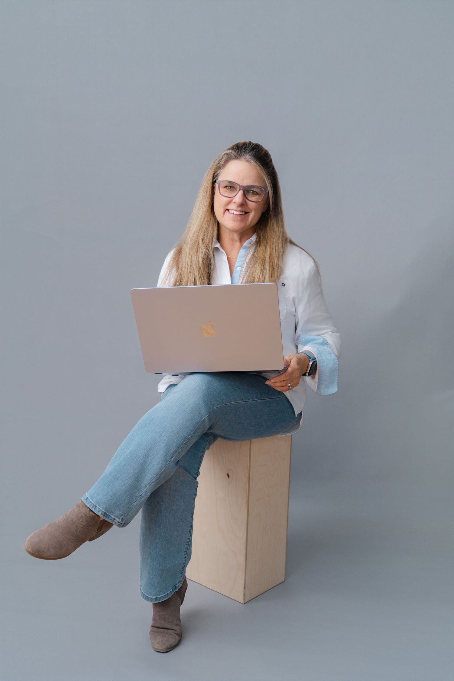 Woman with glasses sitting on a box, smiling while using a laptop, wearing white shirt and jeans.