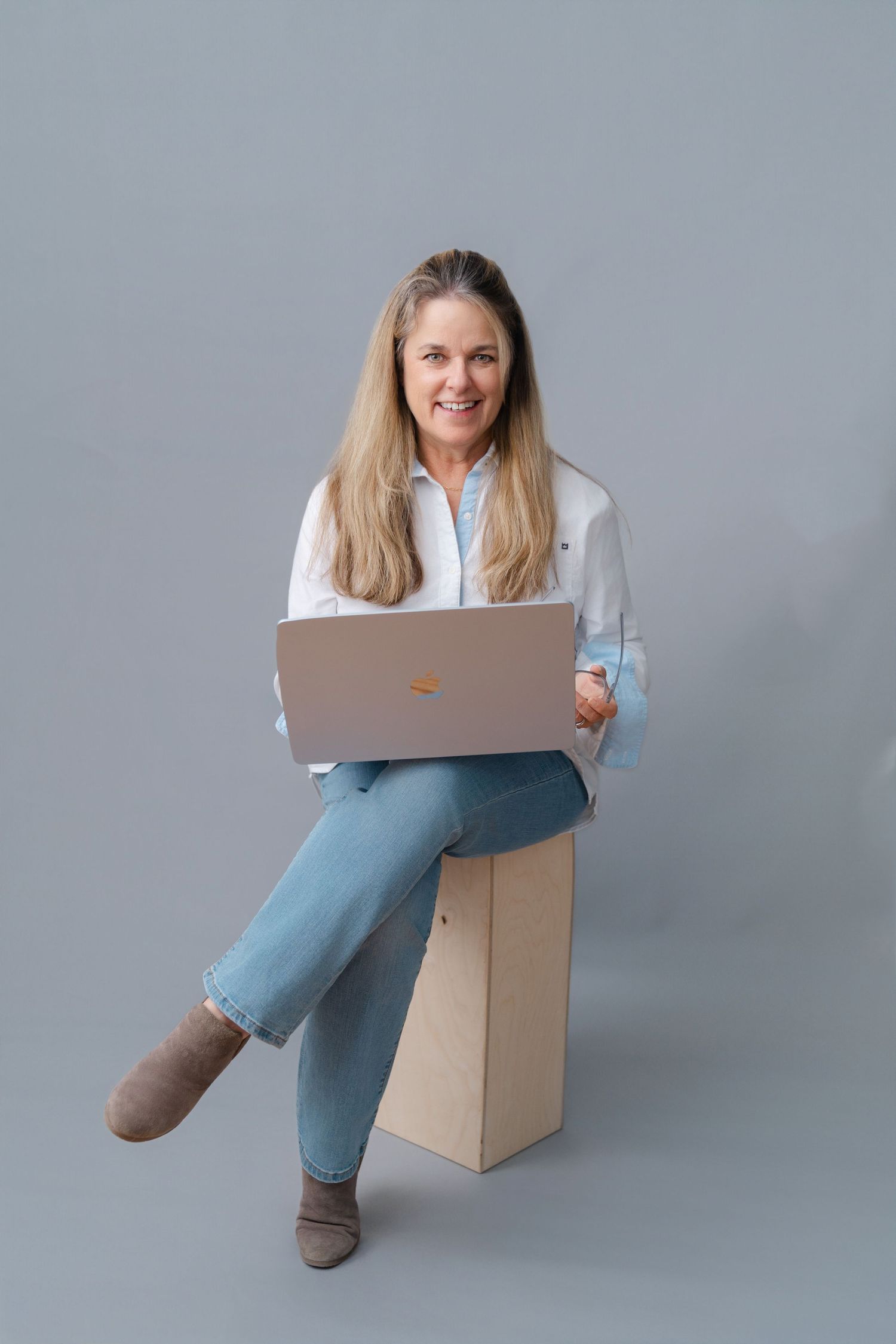 Professional sitting on cardboard box working on laptop wearing jeans and casual attire against gray background.