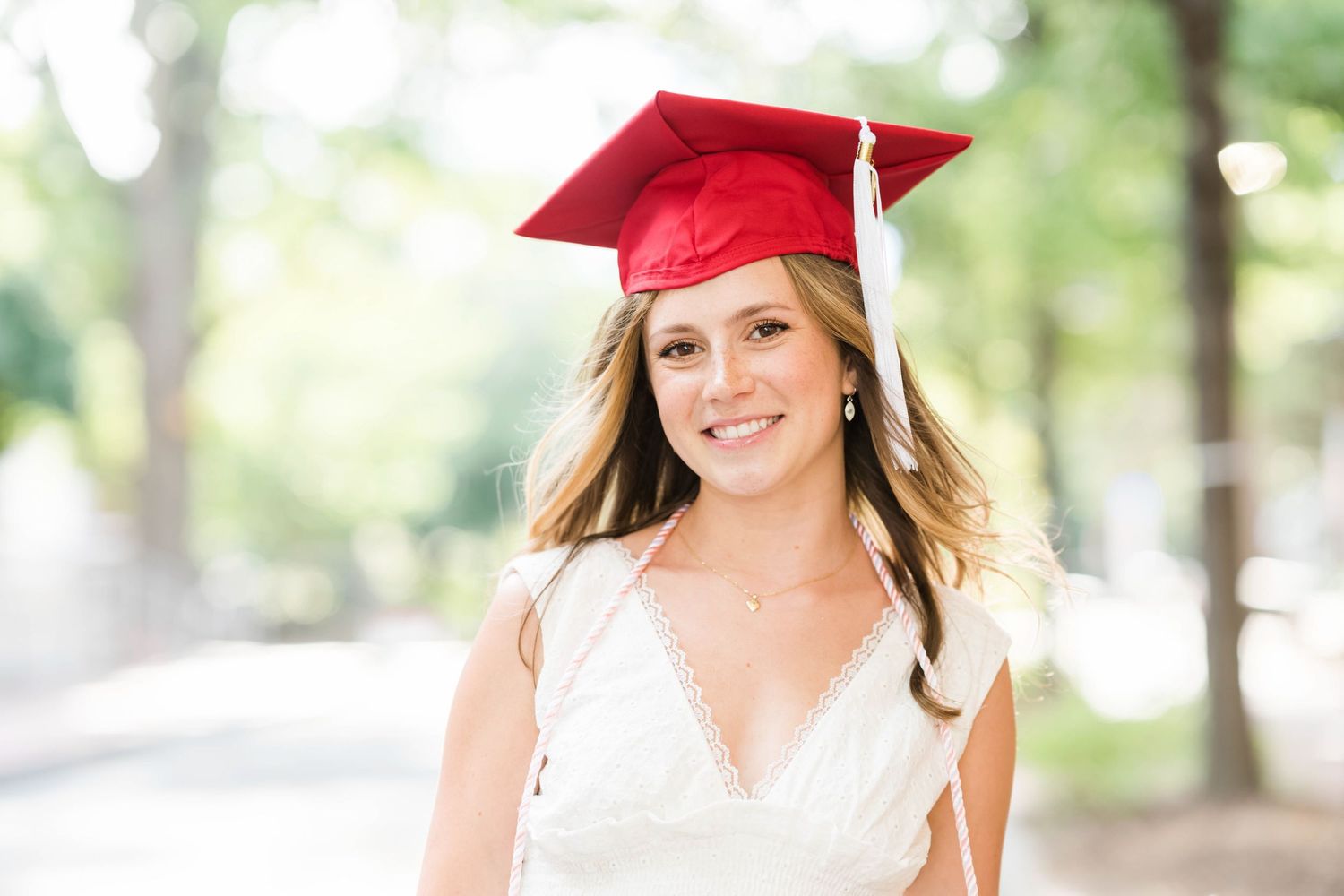 Graduate in red cap and gown smiles brightly during outdoor graduation photo session.