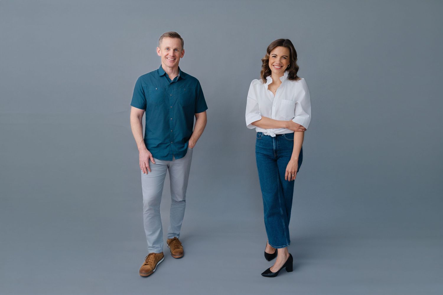 A man and woman stand smiling against a gray background, wearing casual business attire in blue and white tones.