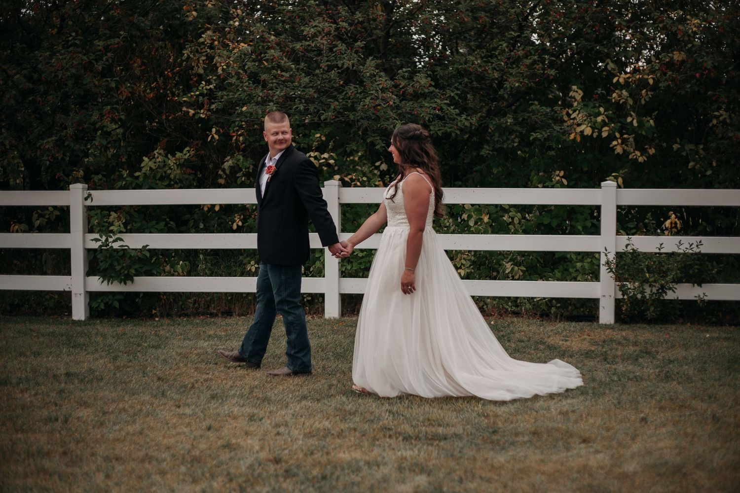 A couple walks together along a white fence on a rustic farm property during their wedding celebration.