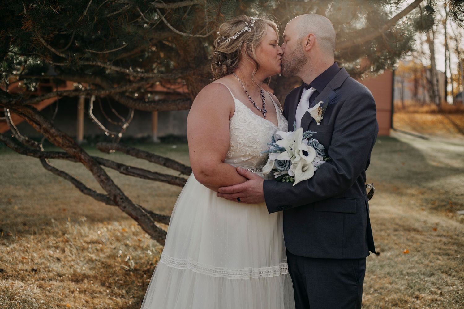 A romantic outdoor wedding moment with a couple embracing while holding a white bouquet at sunset.