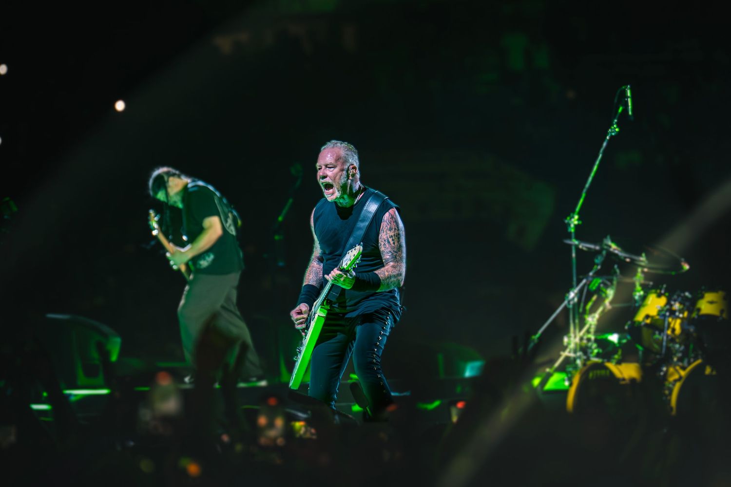 Two rock musicians perform on stage bathed in green light, one playing guitar energetically at a live concert.