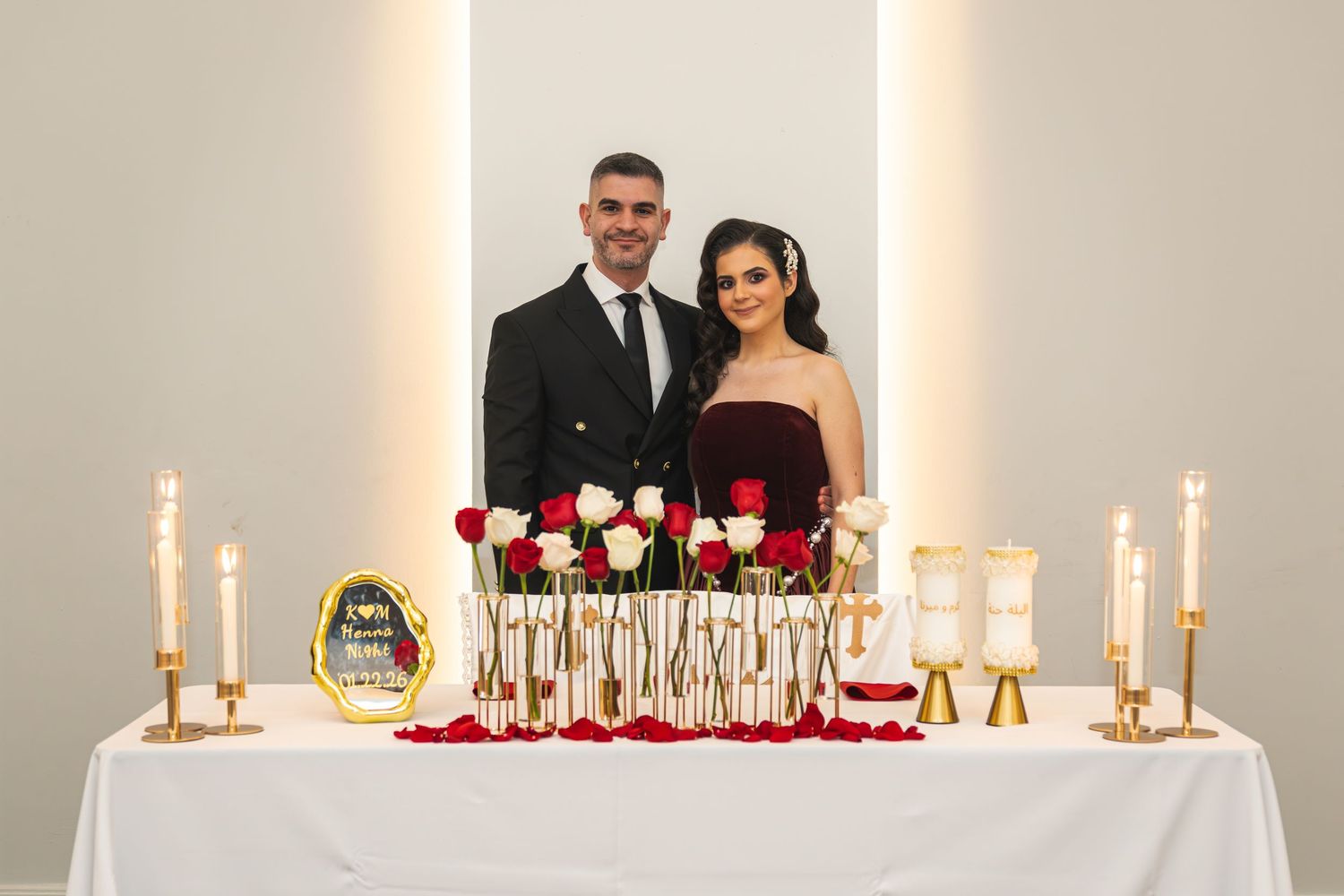 Couple posing at elegant wedding table with red roses, candles, and gold accents against a white backdrop.