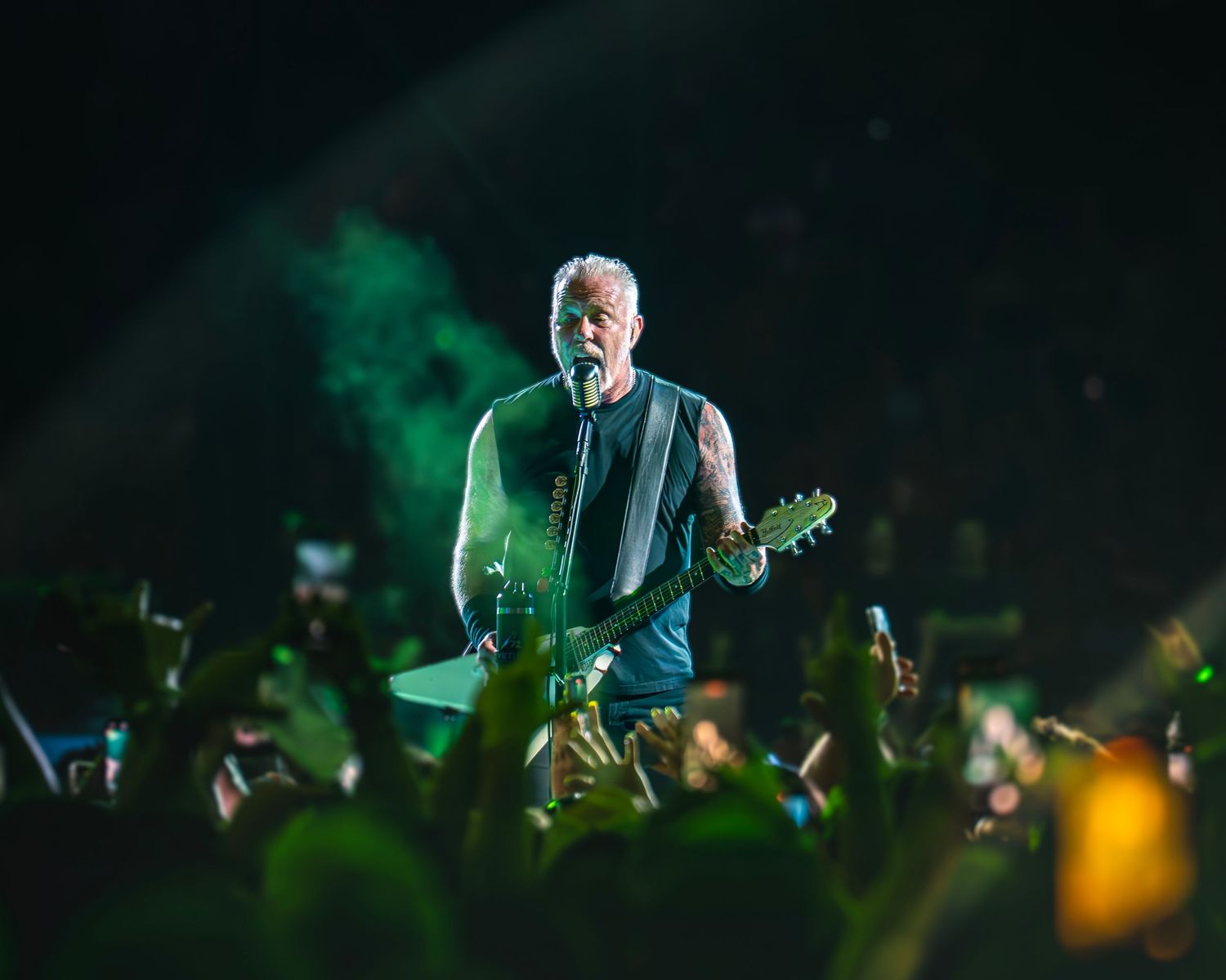 A tattooed rock guitarist performs solo on stage surrounded by green smoke and crowd hands at a live show.