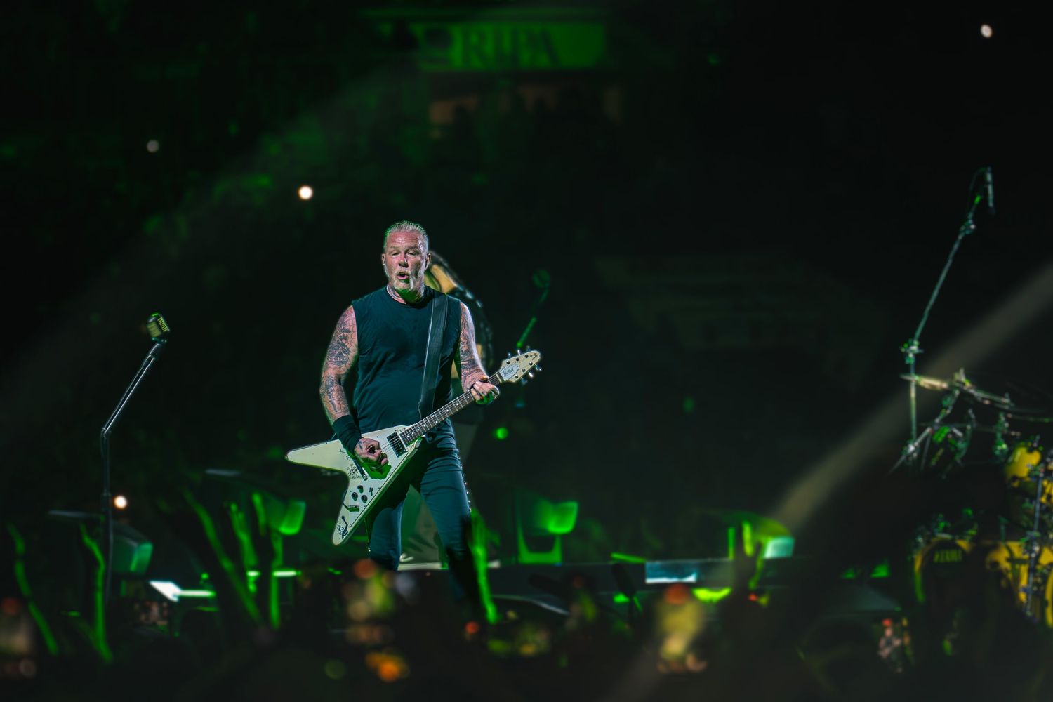 A rock musician plays a white Flying V guitar on a large stage with green spotlights at a live concert.